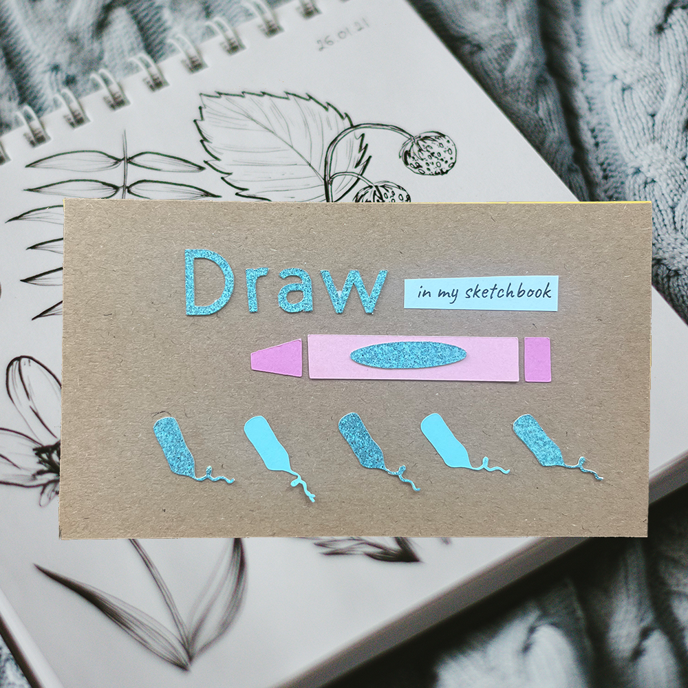 a punch card that reads " draw in my sketchbook" with in blue glitter text. There are 5 pencil cutouts glued on to the card. The card in on top of a picture on a sketchbook