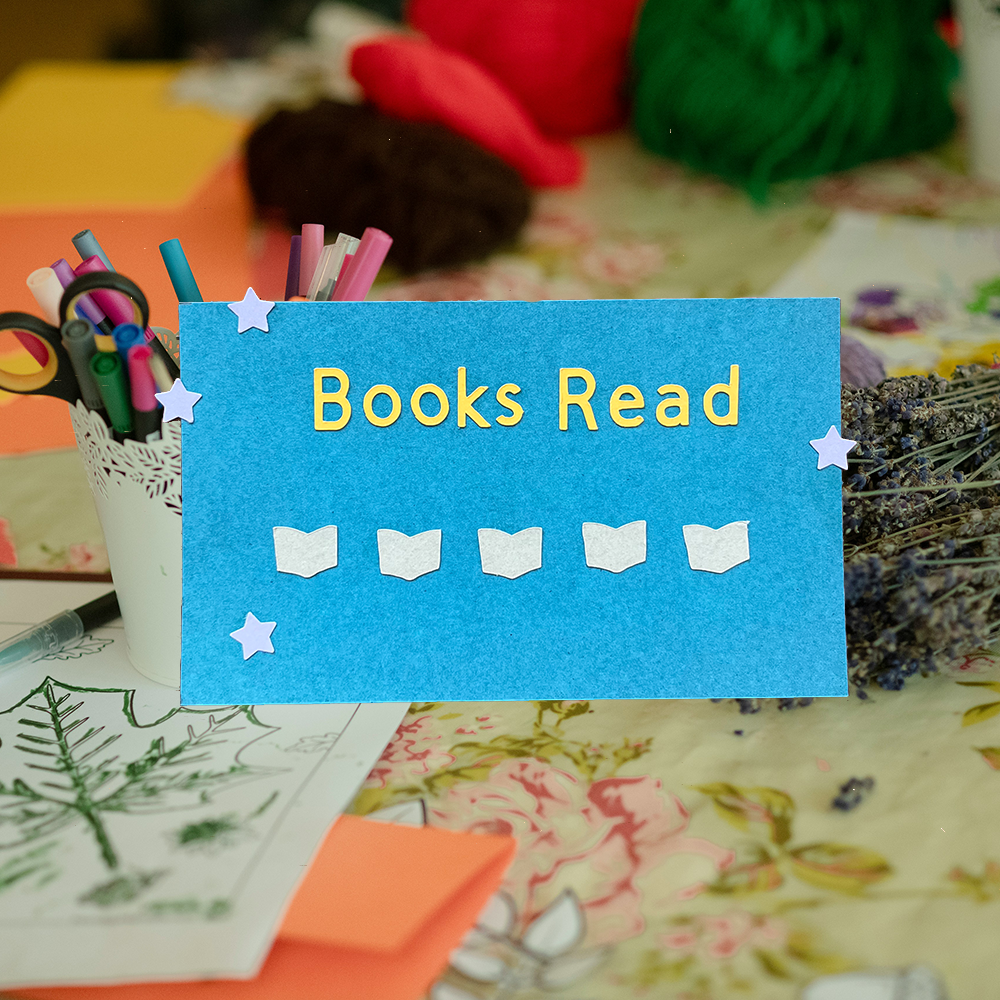 a blue punch card that reads " books read" with 5 tiny book cutouts on the card. The card is on top of a photo of art supplies and dried flowers.