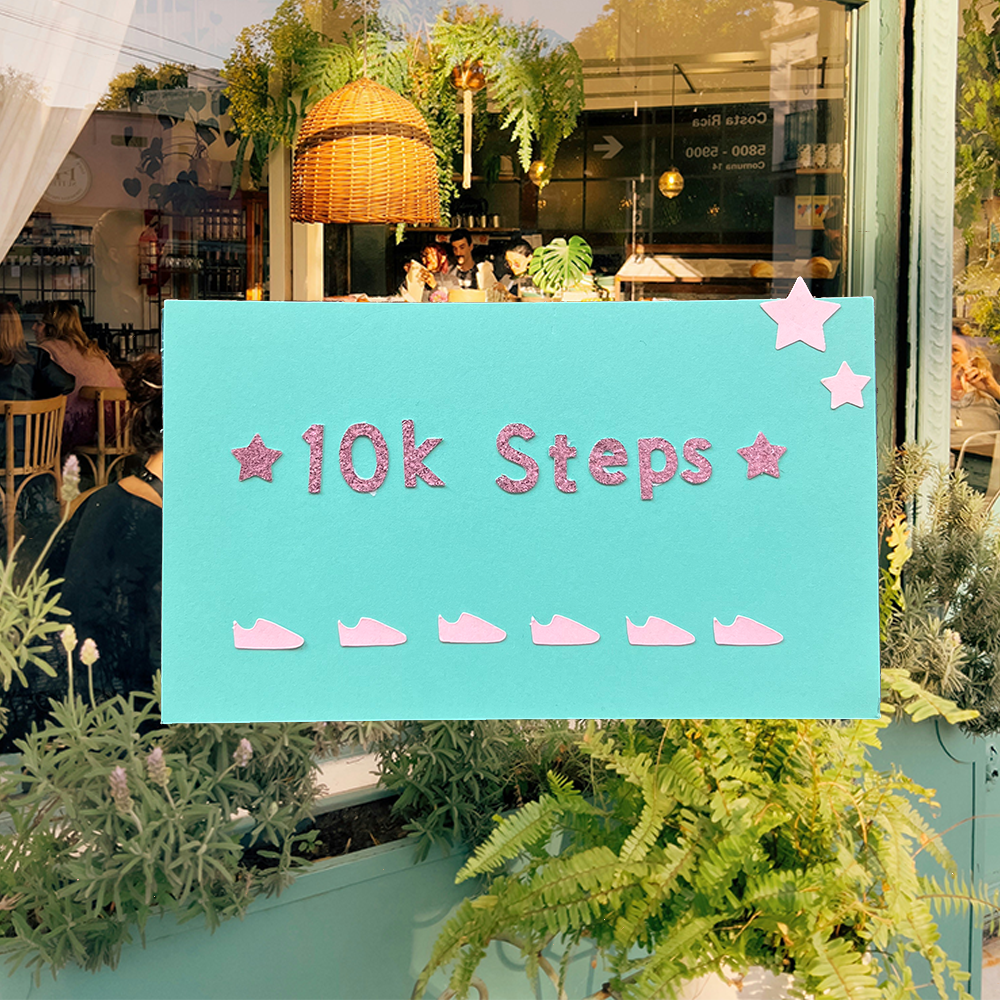 a punch card that reads " 10k steps" in pink glitter letters. The card has 6 pink shoes next to each other to punch. The punch card is on top of a picture of a coffee shop.