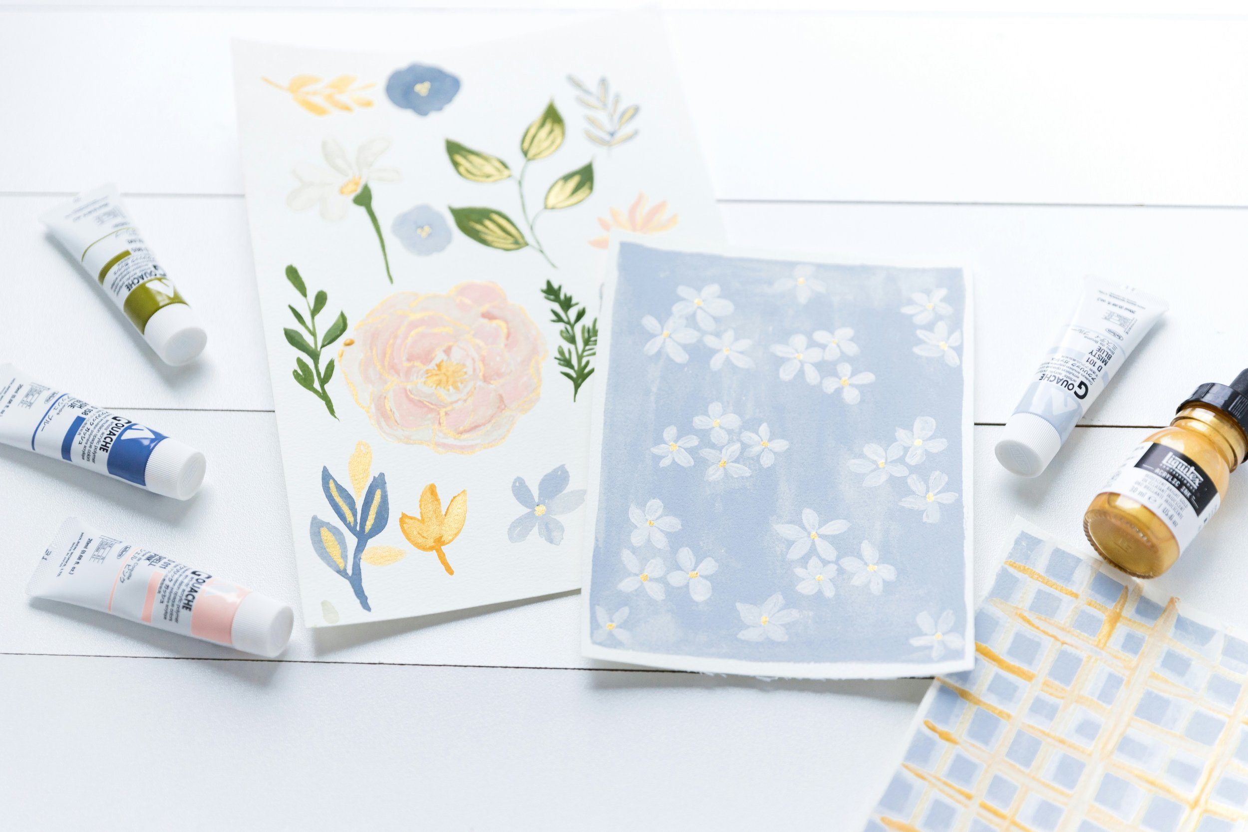 paint tubes and three watercolor paintings laying on a white table