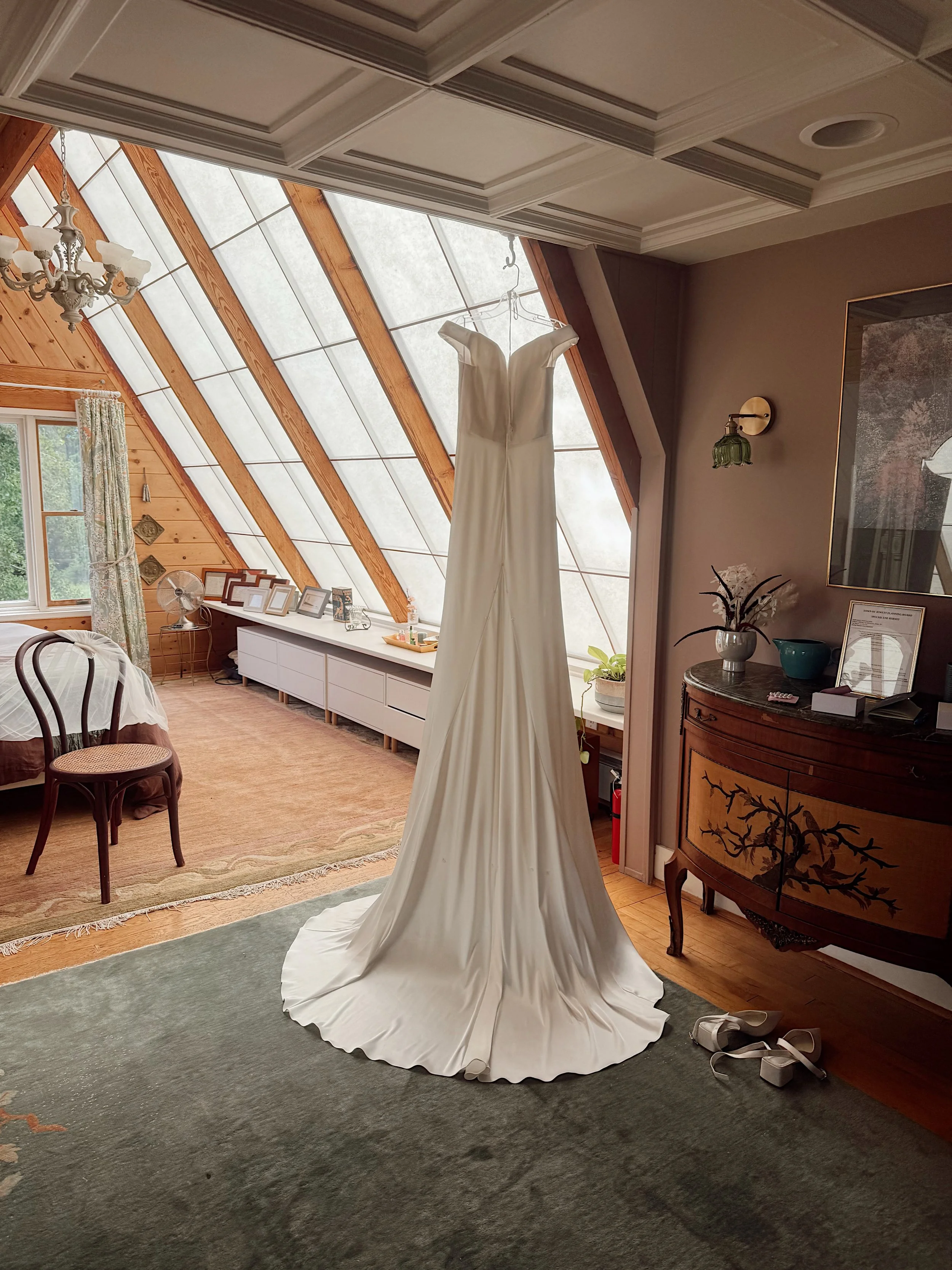 A wedding dress hanging in a room with a vaulted ceiling and large skylight windows, with shoes on the floor nearby.