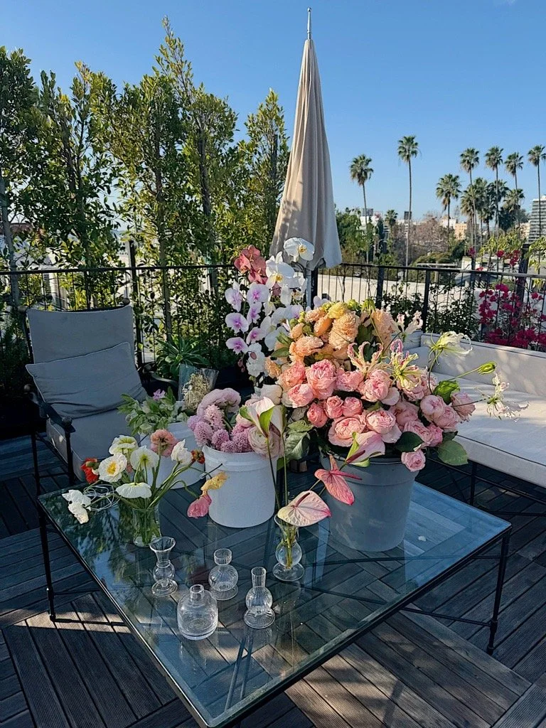 Outdoor patio table with pink and white flowers in white and light blue pots, surrounded by chairs, a closed umbrella, and greenery with palm trees in the background.