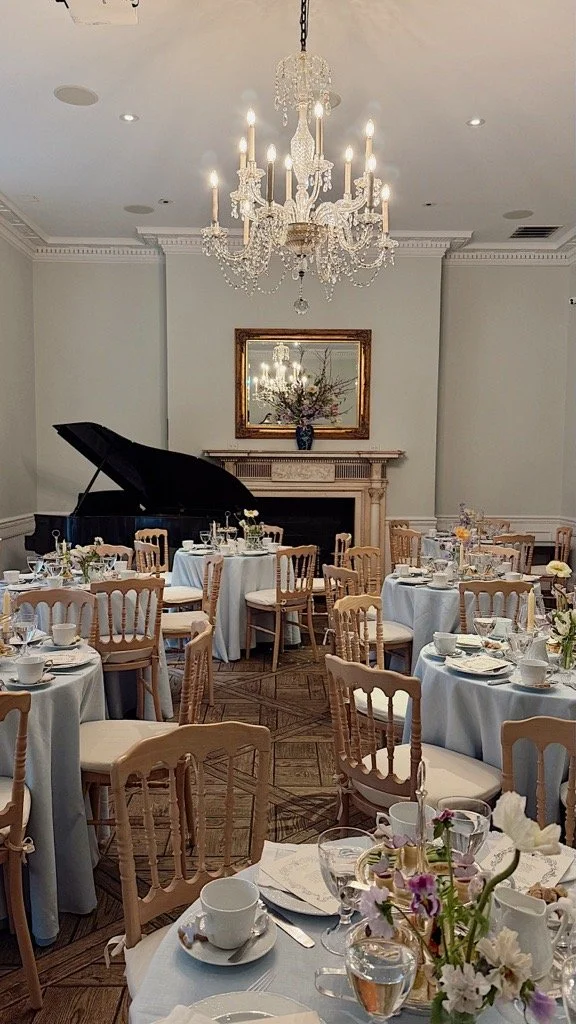 Elegant banquet room decorated for a formal event with round tables draped in white tablecloths, set with fine china, silverware, glasses, and floral centerpieces. A grand chandelier hangs from the ceiling, and a black grand piano is positioned in fr