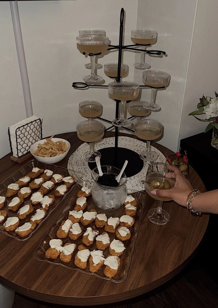 Cocktail glasses with greenish drinks on a tiered stand, bowl of French fries, and plates of chicken nuggets with white dipping sauce on a wooden table.