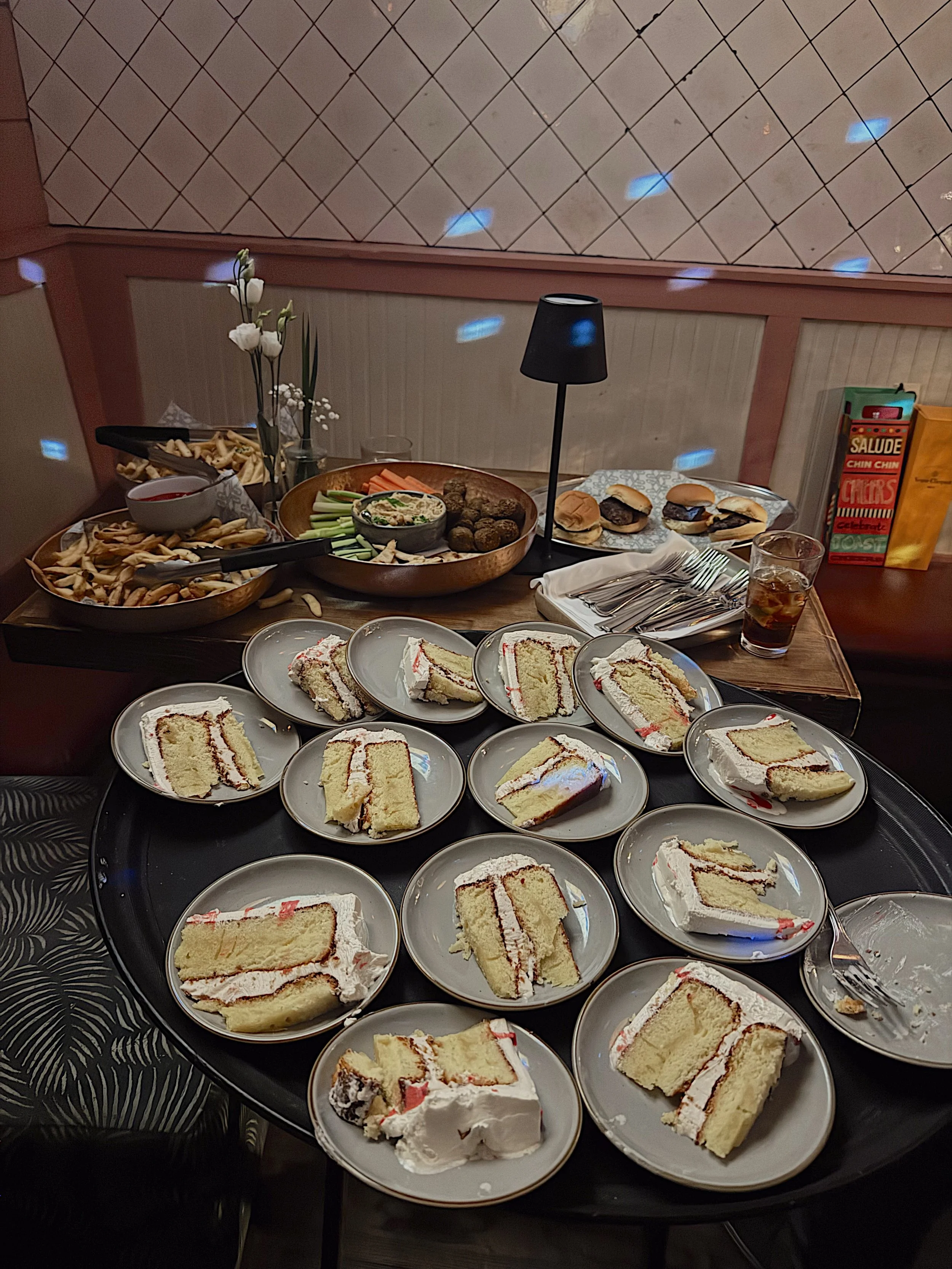 A table setting with multiple plates of sliced cake, including pieces with white frosting and red jam or filling, along with various desserts such as bread rolls, and a tray of snacks. The table also has a drinks glass with a dark beverage, utensils,