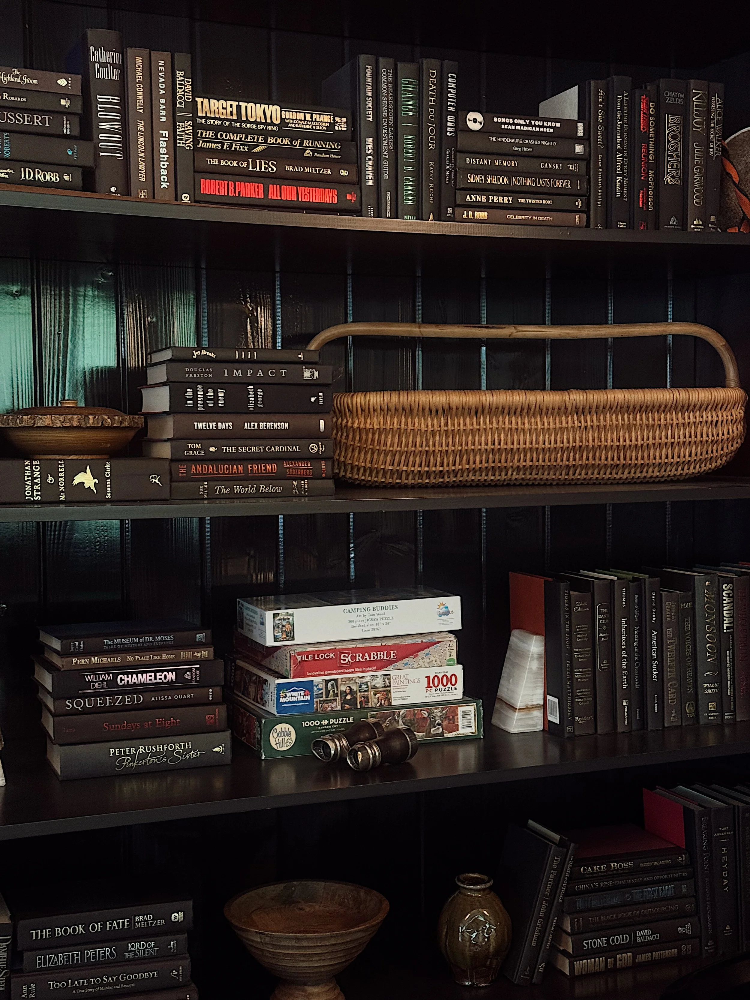 A bookshelf containing multiple stacks of books, with some books stacked vertically and others horizontally, a woven basket, a wooden bowl, a small glass bottle, and a pair of binoculars placed on the shelf.