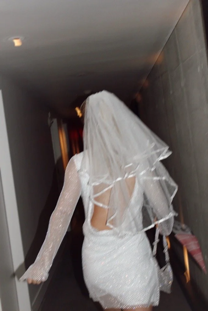 Woman in a sparkly dress and veil walking down a hallway, with her back to the camera.