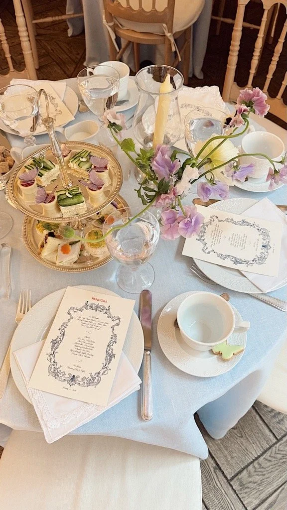 Elegant dining table set with tea cups, wine glasses, a gold tiered serving tray, pastel pink and purple flowers, candles, and printed menus.