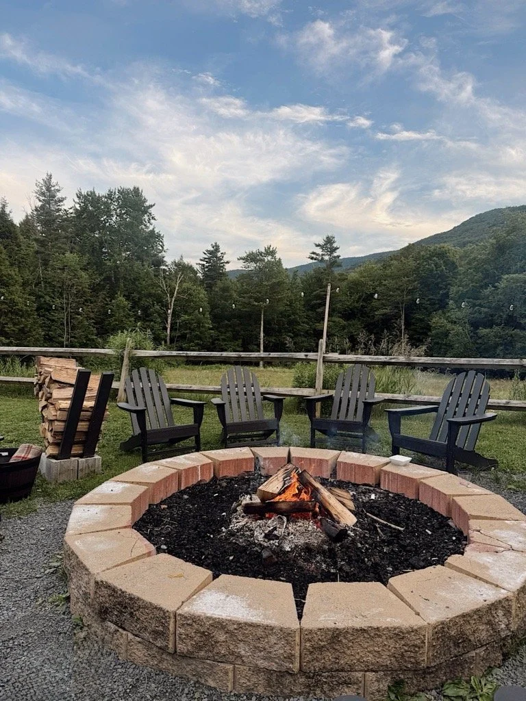 An outdoor fire pit with burning logs surrounded by four Adirondack chairs, set on a grassy area with trees and a mountain in the background.