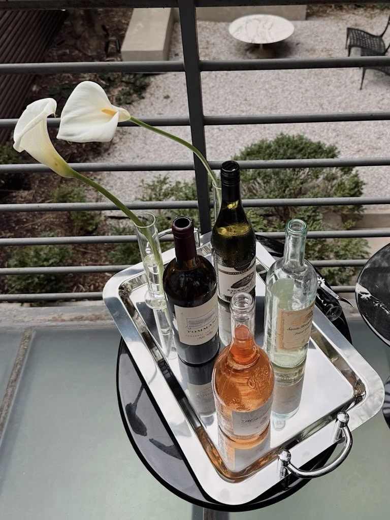 A tray with four bottles of wine and a glass vase with white calla lilies, on a balcony with outdoor furniture and a garden below.
