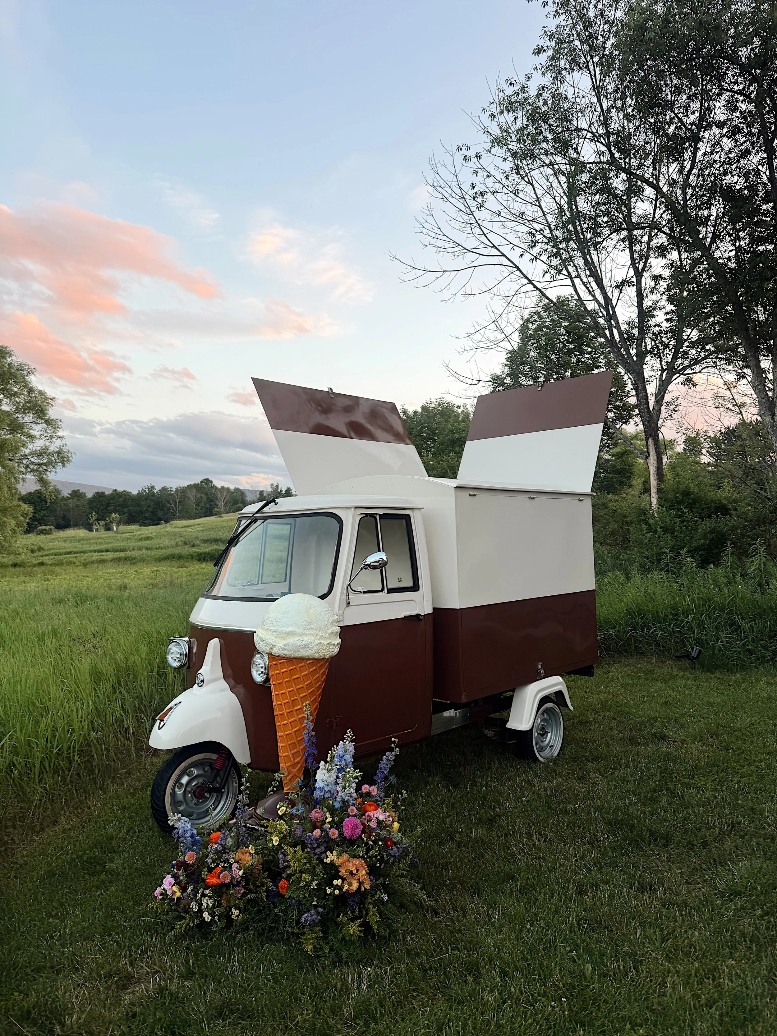A small three-wheeled ice cream cart decorated with flowers in front of a grassy field and trees during sunset.