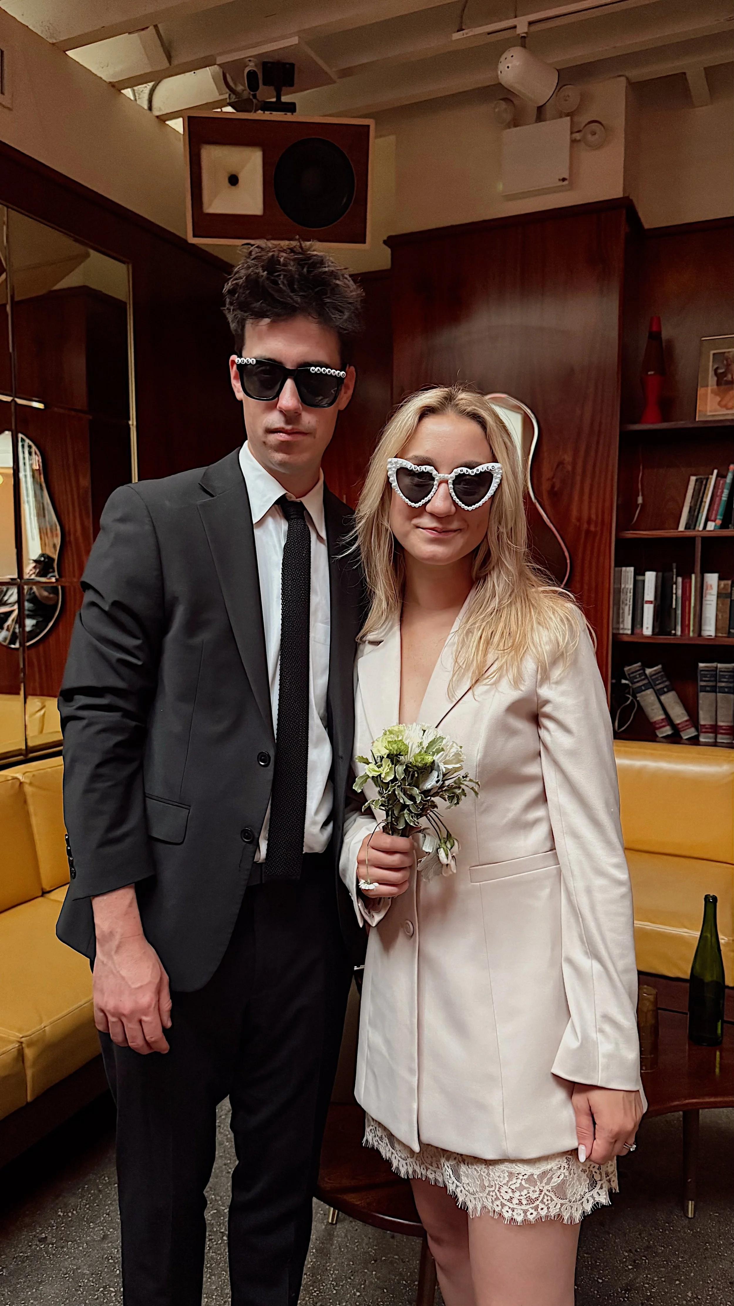 A man and woman dressed in formal attire, both wearing sunglasses, standing close together indoors. The woman is holding a small bouquet of flowers. The background features wooden paneling, bookshelves, a yellow couch, and decorative items.