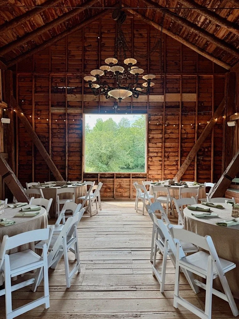 Interior of a rustic barn with wooden walls and ceiling, decorated for a gathering with round tables covered in beige tablecloths, white chairs, and a large black chandelier hanging from the ceiling. A large window at the front views green trees outs