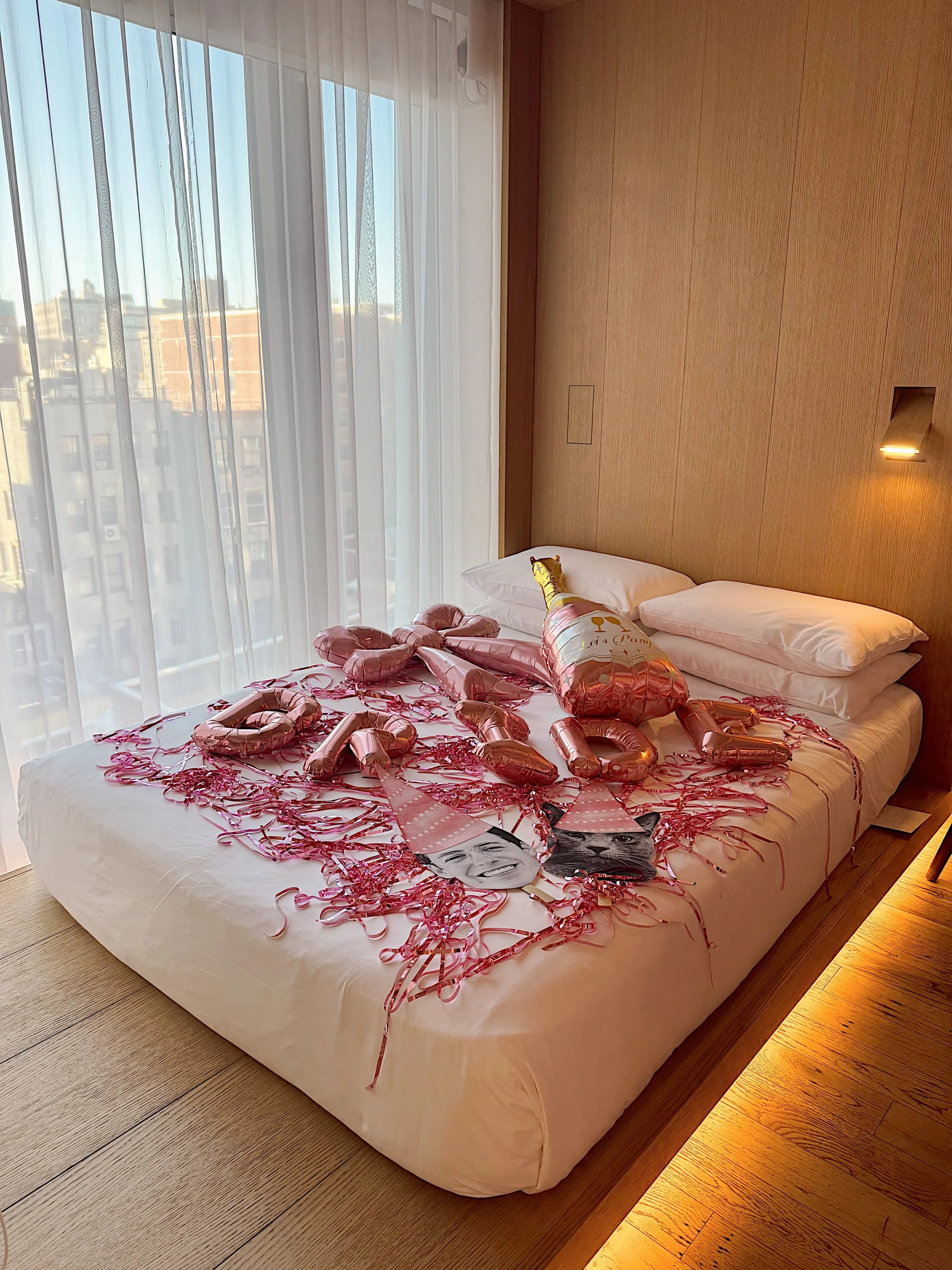 Hotel room with a bed decorated with pink foil balloons, pink and red confetti, and paper photos of a smiling woman and a dog, possibly for a celebration.