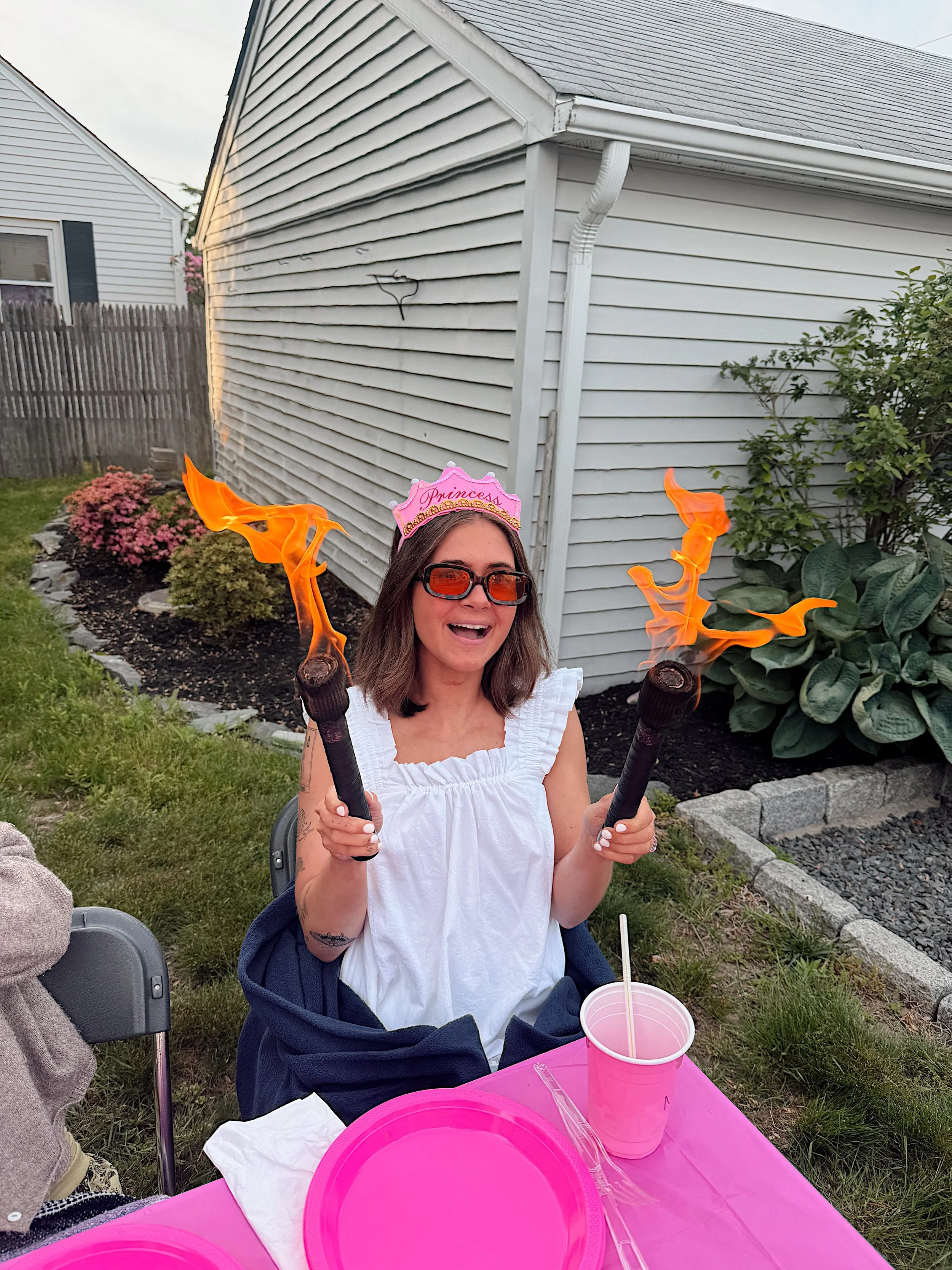 Woman wearing a pink princess crown and sunglasses, holding flaming torches, sitting outdoors next to a pink table with pink cups and containers, on a lawn with houses and bushes in the background.
