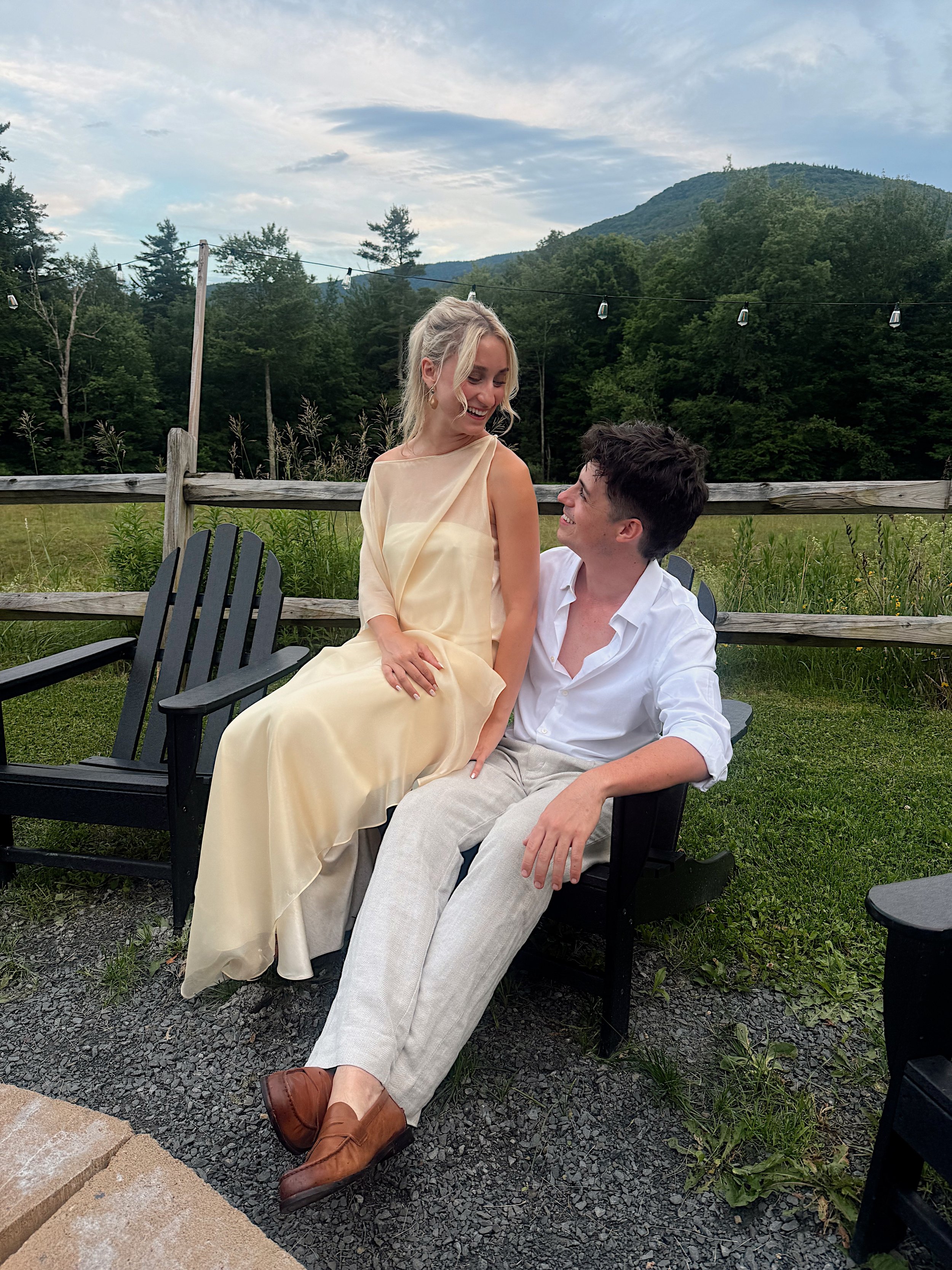A man and woman sitting together outdoors on black chairs in a grassy area, smiling and looking at each other, with a fence, trees, and mountains in the background.