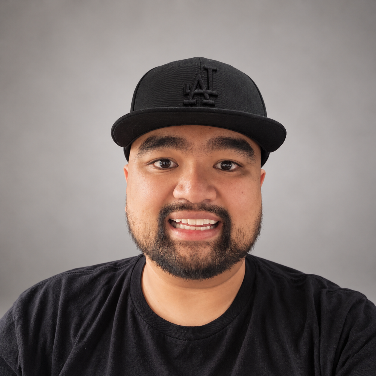 A man with a cap and a beard, smiling slightly while looking off to the side. He is wearing a dark sweater and is seated indoors with a blurred plant in the background.