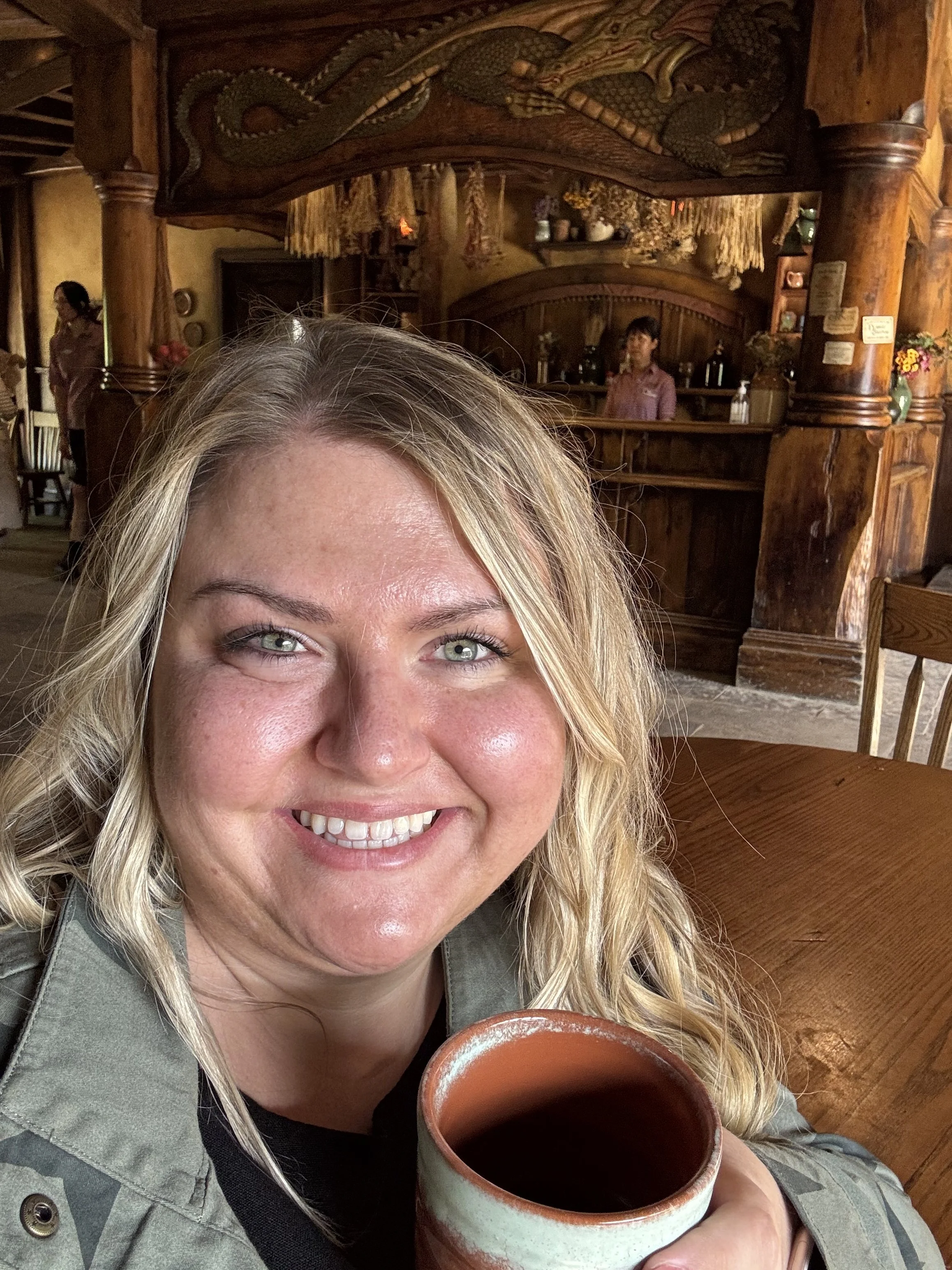 A woman smiling while holding a mug of coffee or tea inside a rustic wooden cafe or restaurant.