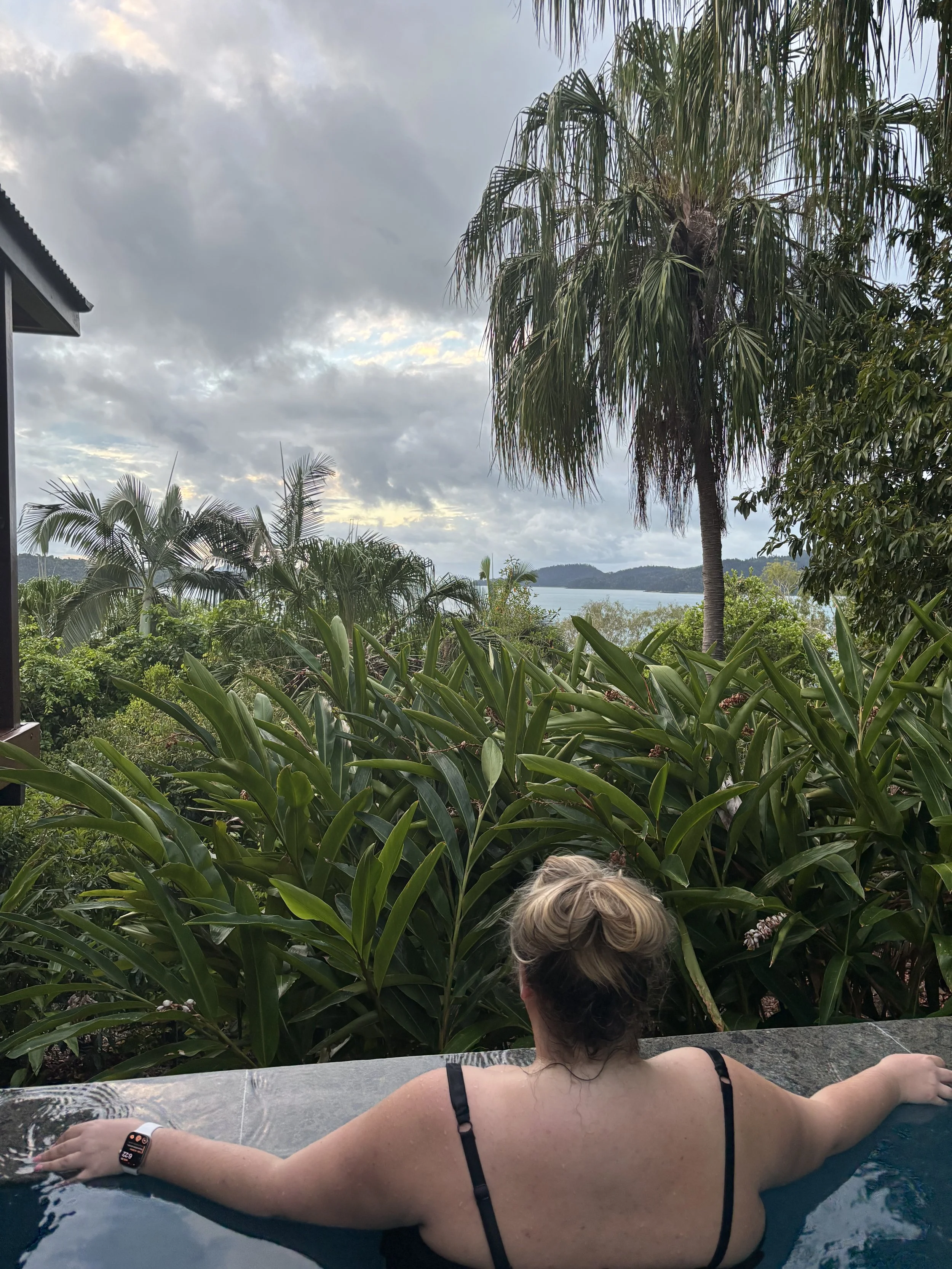 A woman with blonde hair tied back, wearing a black tank top and a fitness tracker on her wrist, is relaxing in a hot tub with her arms stretched out on the edge. She is surrounded by lush green tropical plants, including a palm tree, with a cloudy sky and distant water body in the background.