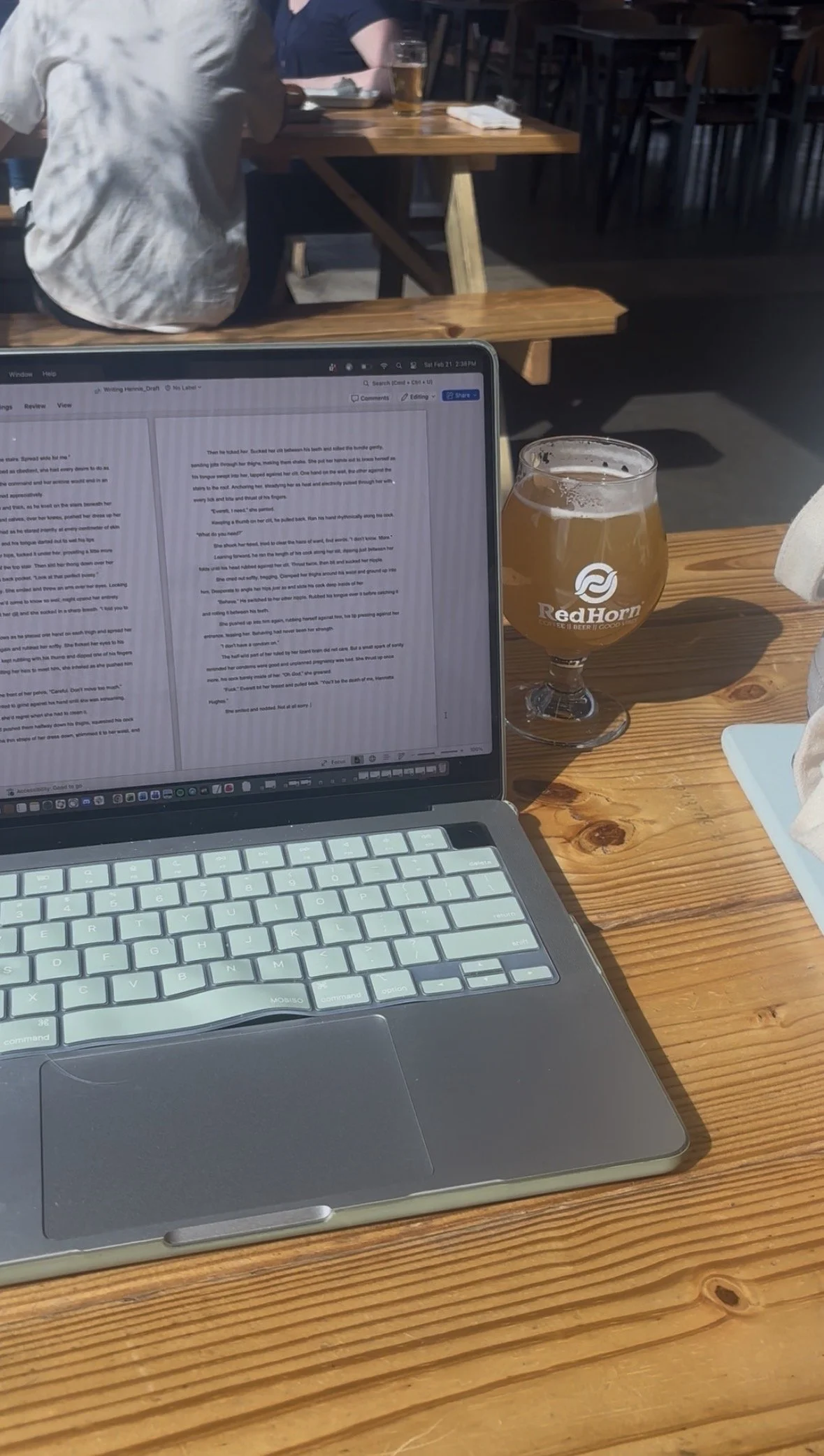 A laptop on a wooden table with an open document on the screen, a glass of beer labeled 'RedHorn', and another person in the background at a different table with a beer.