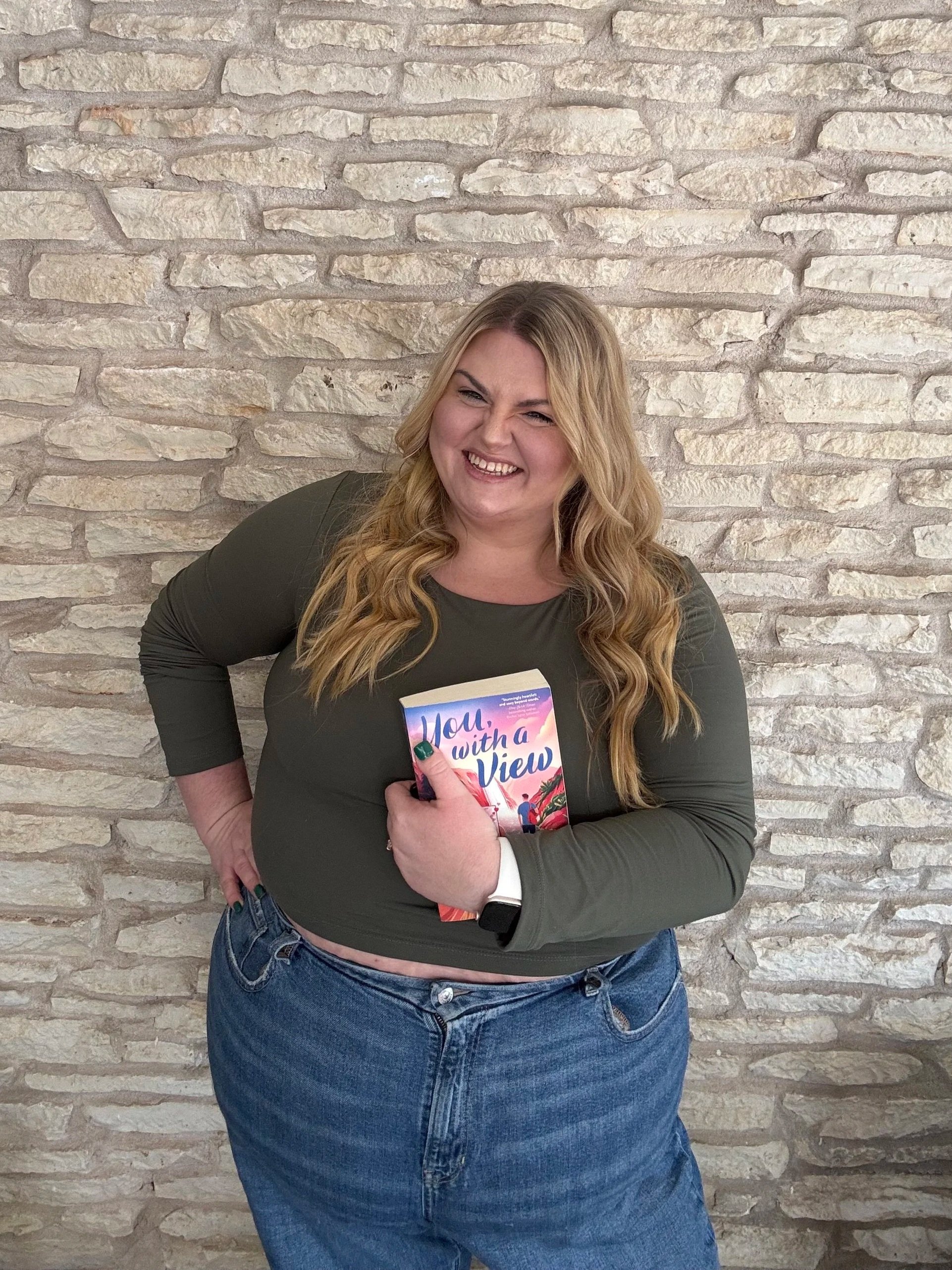 A woman with long, wavy blonde hair, wearing a dark green long-sleeve shirt and blue jeans, holding a colorful book titled "You, with a View" and smiling in front of a beige stone wall.