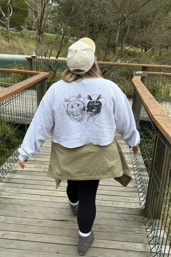 Person walking on a wooden pathway with a hoodie featuring animal portraits, surrounded by trees and greenery.