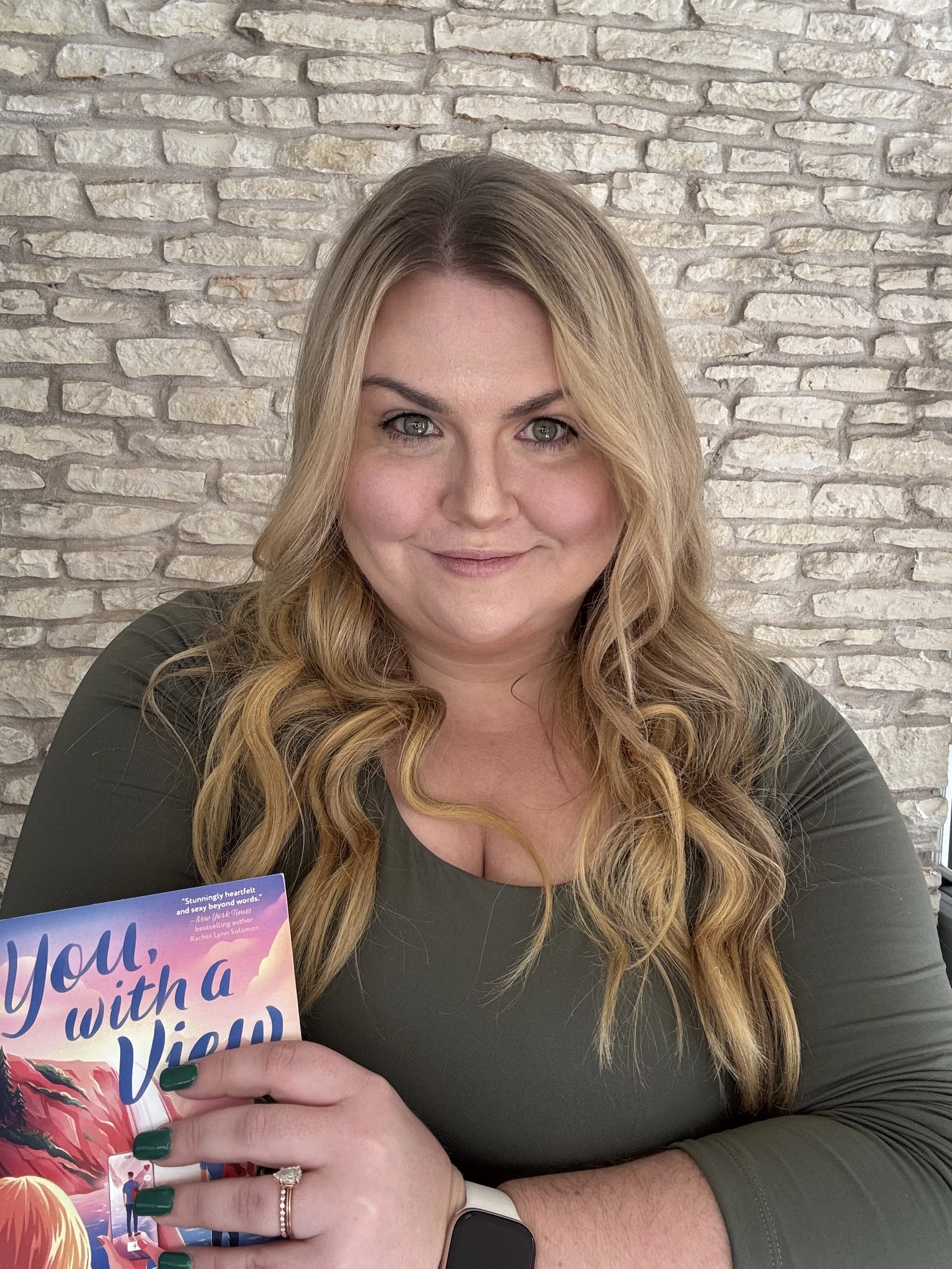 A woman with long wavy hair, wearing a green top, holding a book titled 'You, with a View'. She is seated in front of a stone wall. She has a ring and a bracelet on her left hand, and a smartwatch on her right wrist.