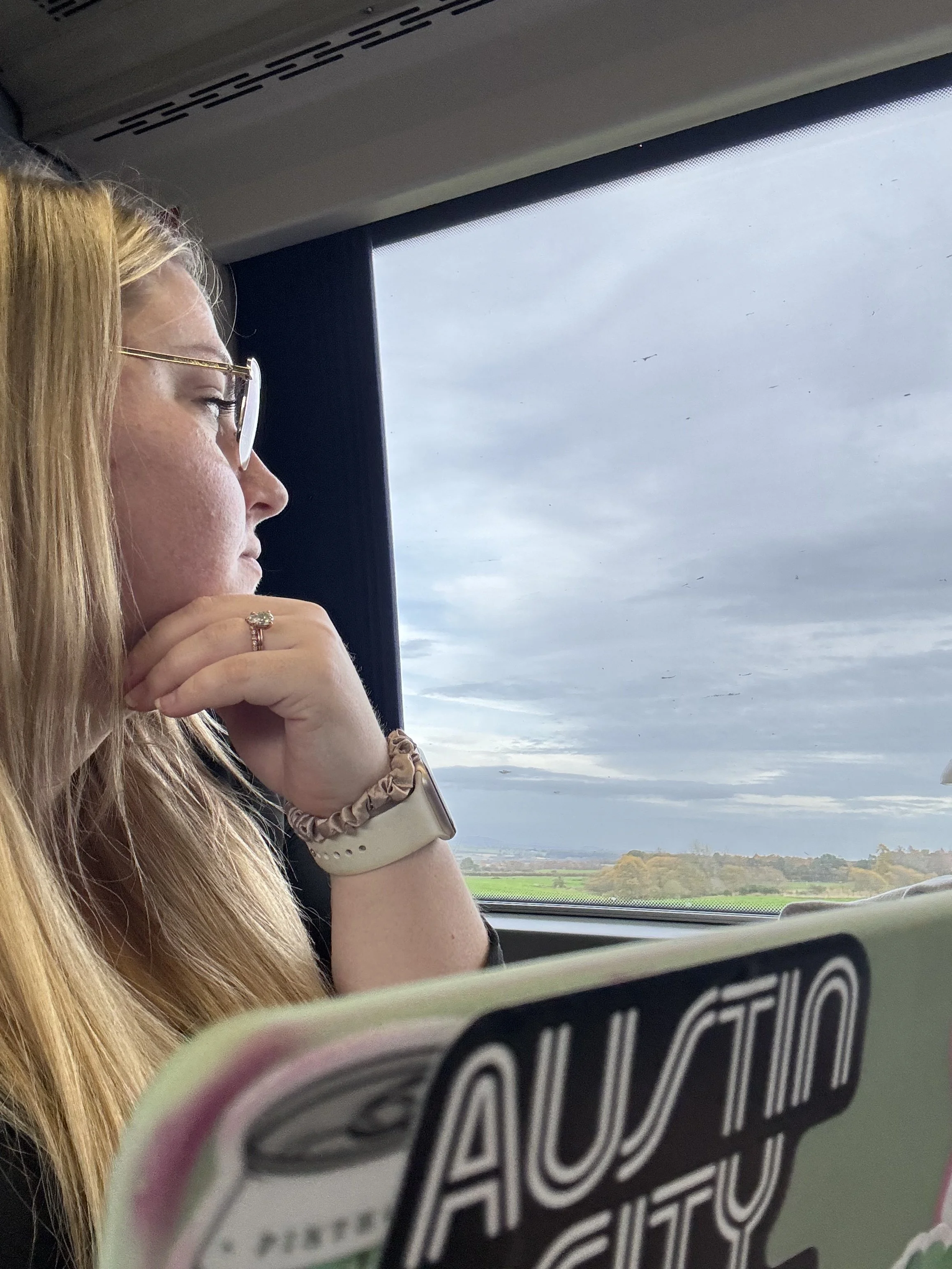 A woman with glasses and long blonde hair reading a book titled 'Atomic City' on a bus. She has a ring on her finger, a bracelet, and a wristwatch. The background shows cloudy skies and a landscape with trees and fields seen through the bus window.