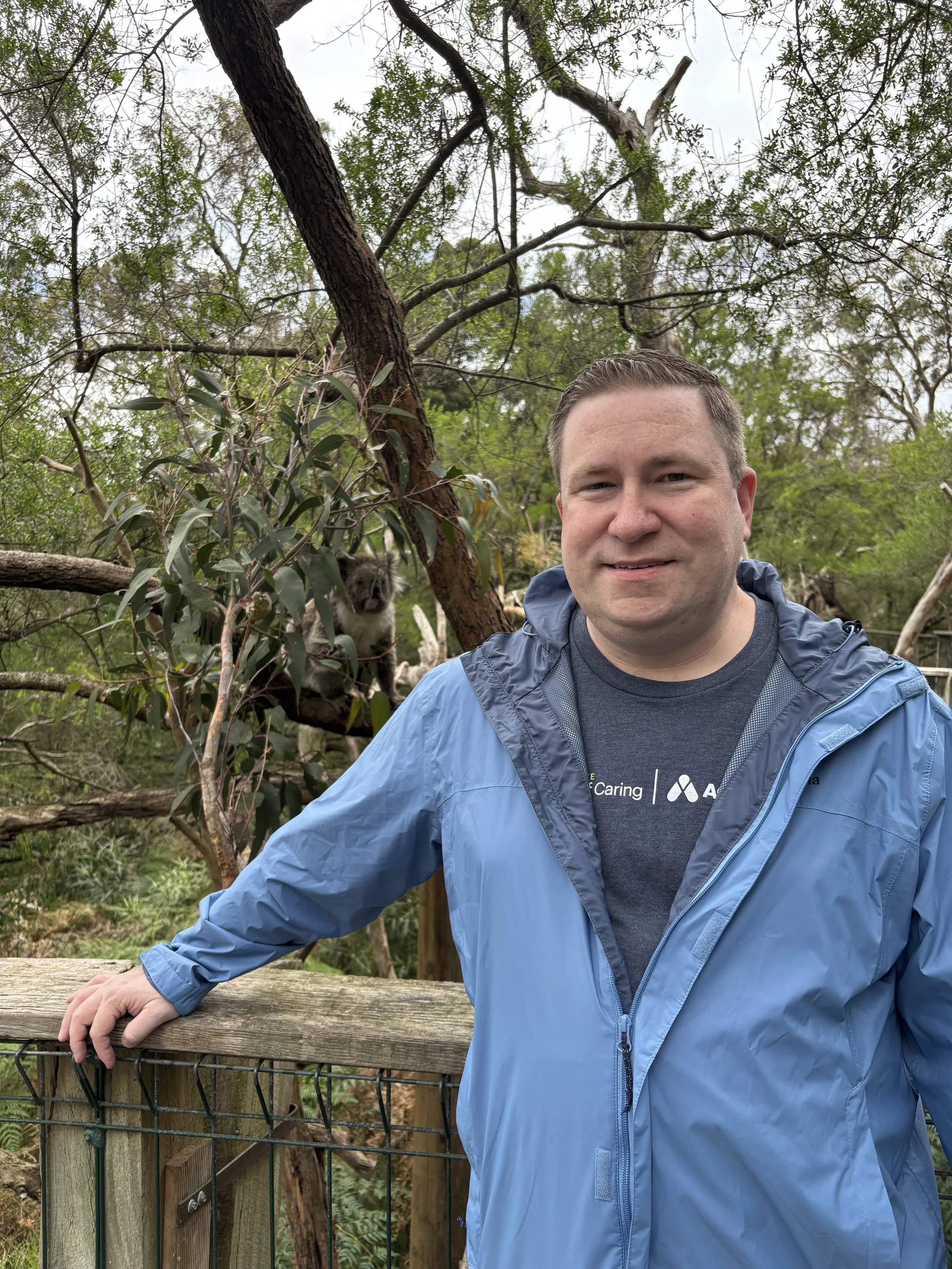 A smiling man wearing a blue jacket standing near a wooden fence in a forested area with trees and greenery, holding onto the fence with one hand.