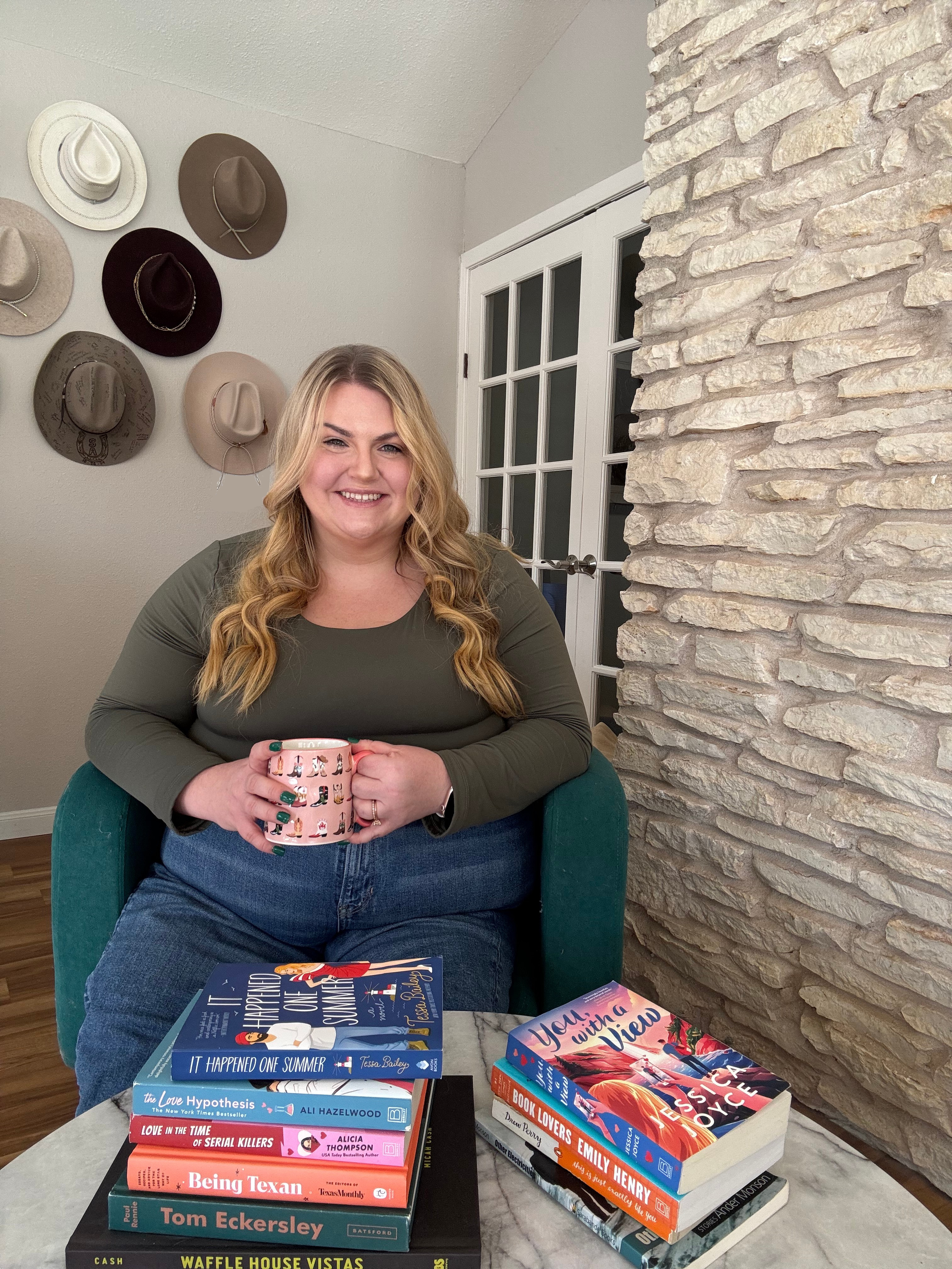 A woman sitting at a table holding a pink mug, with a smile. In front of her are two stacks of books on a marble table, and a book on her lap.