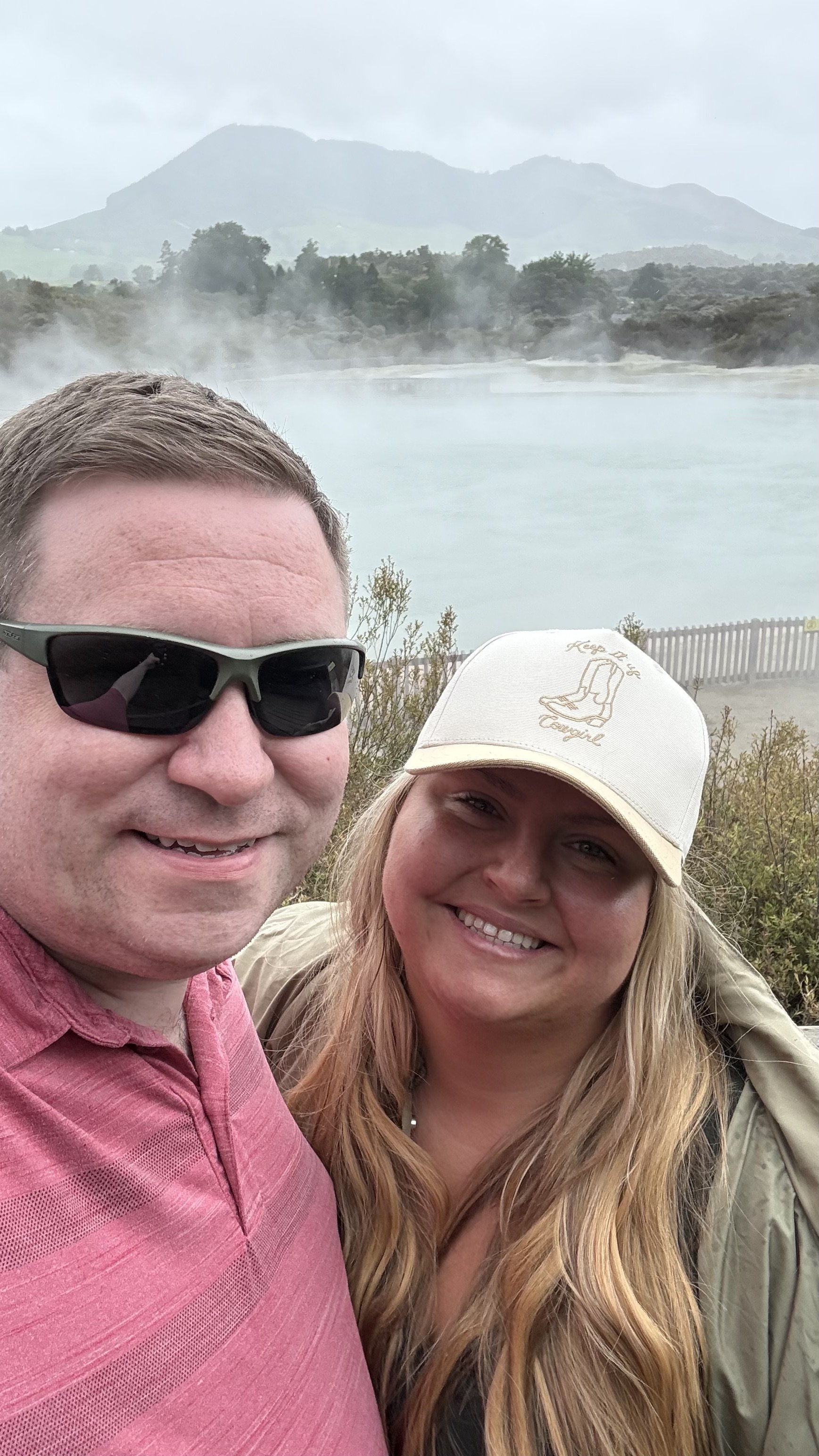 A man and woman taking a selfie outdoors with a steam-covered hot spring and a mountain in the background.