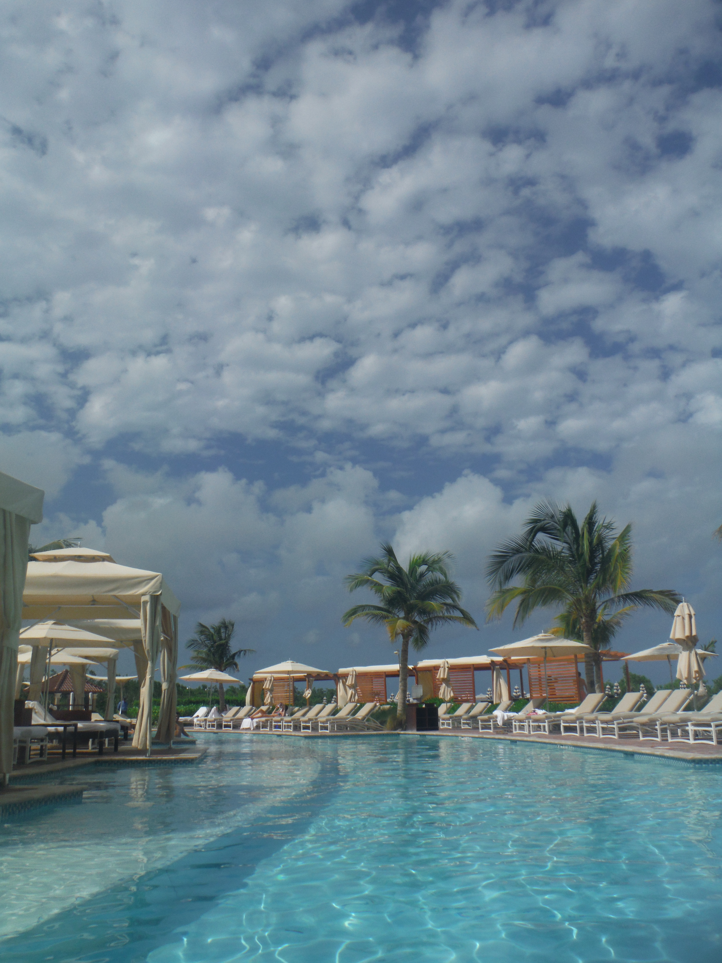 A swimming pool area with lounge chairs and umbrellas, palm trees, and a partly cloudy sky.