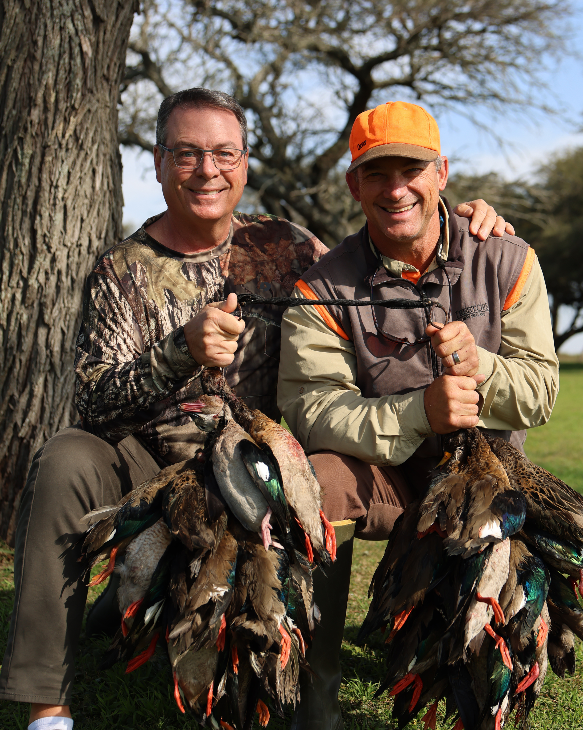 Two men smiling and posing outdoors, holding a large group of dead pheasants, with trees and blue sky in the background.
