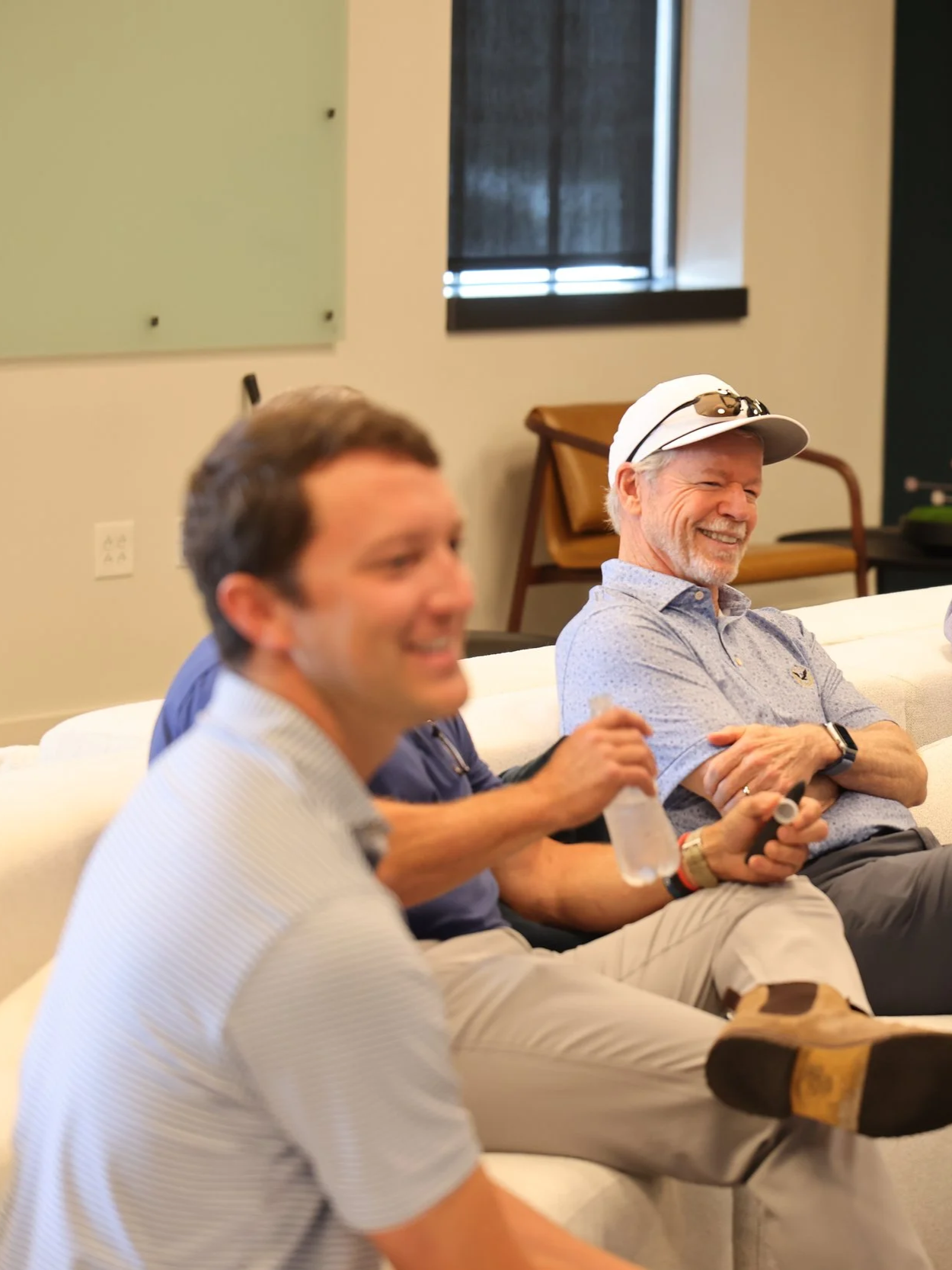 Three men sitting on a couch, smiling and laughing during a meeting or event in a room with a window and wall chalkboards.