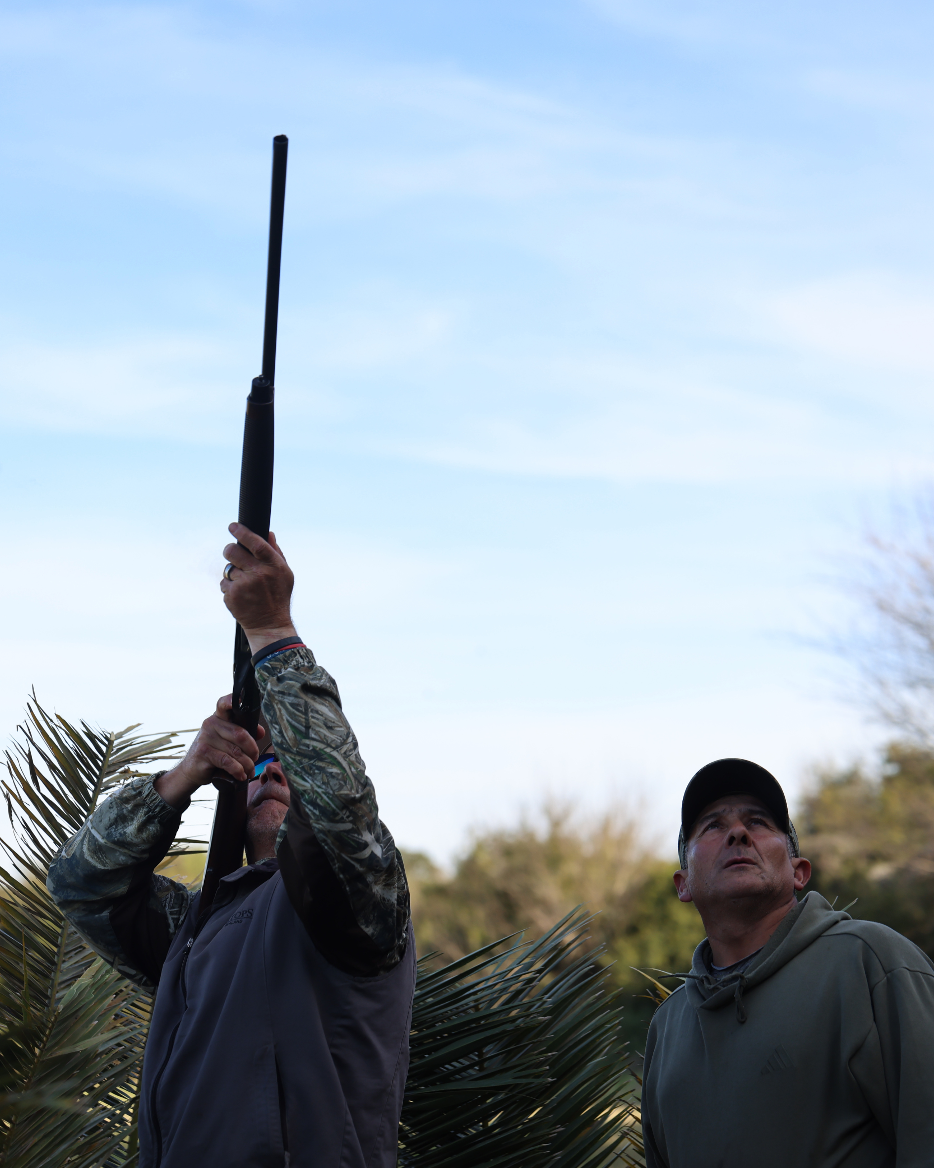 Two men outdoors near palm trees, one holding a black pole or tool up towards the sky, while the other looks on. The sky is mostly clear with some wispy clouds.