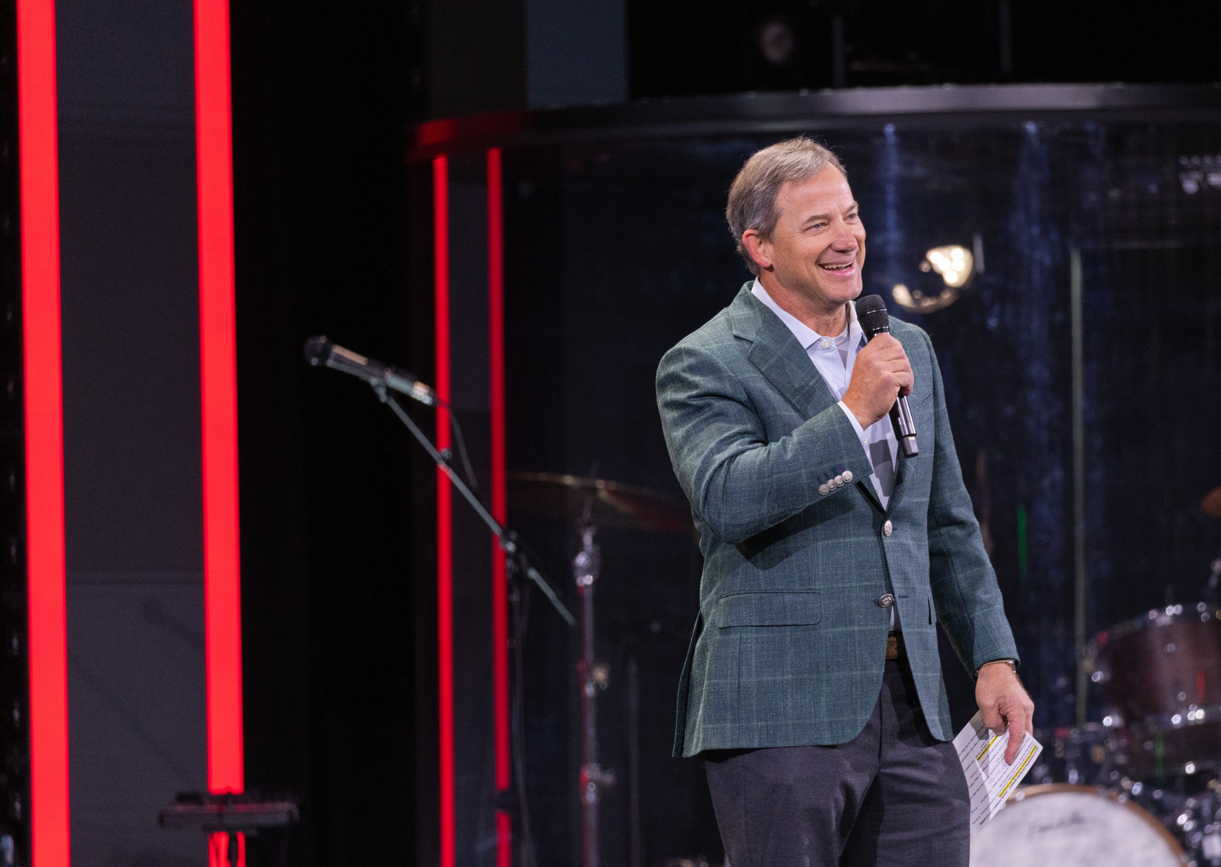 A man in a green plaid blazer holding a microphone and gesture, standing on a stage with a black background and red stage lighting.