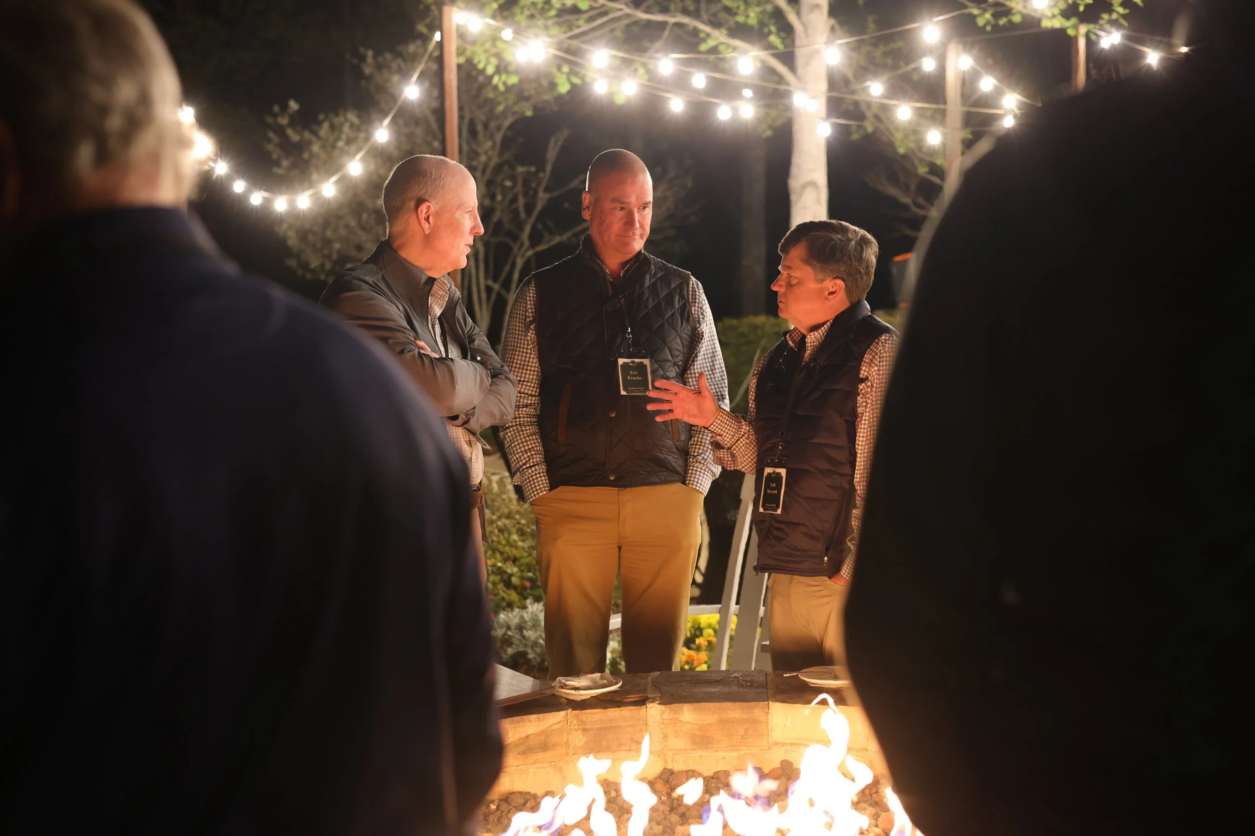 People gathered around a fire pit at night outdoors, with string lights hanging above, engaged in conversation.