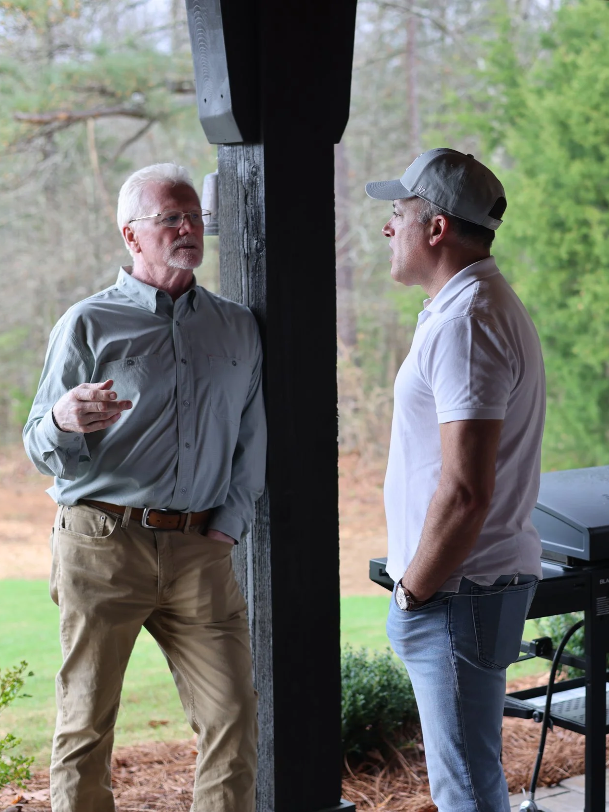 Two men are having a conversation outdoors under a wooden structure, with trees and greenery in the background. One man wears a light gray button-up shirt and khaki pants, while the other wears a white polo shirt, jeans, a cap, and a watch.