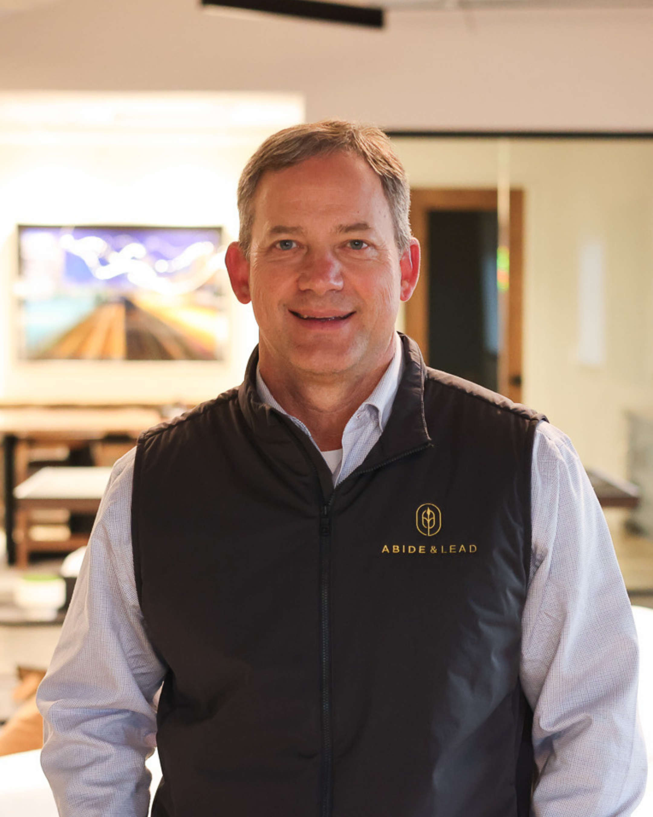 Man smiling in indoor setting, wearing a black vest with 'ABIDE & LEAD' logo over a white shirt, with blurred background of artwork and furniture.