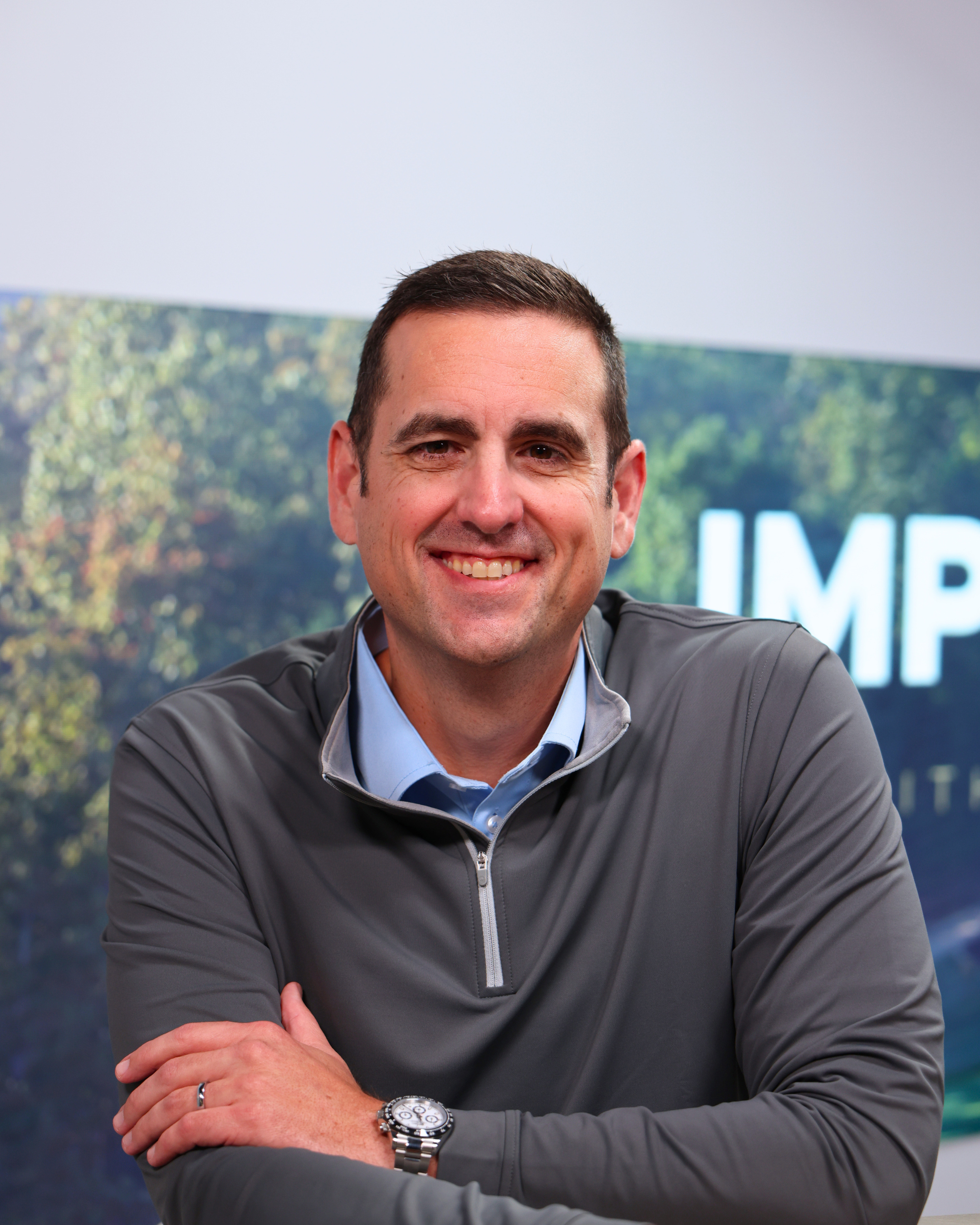 A smiling man with short brown hair, wearing a gray jacket over a blue collared shirt, sitting with arms crossed and wearing a silver wristwatch, in front of a backdrop with trees and a large sign or display.