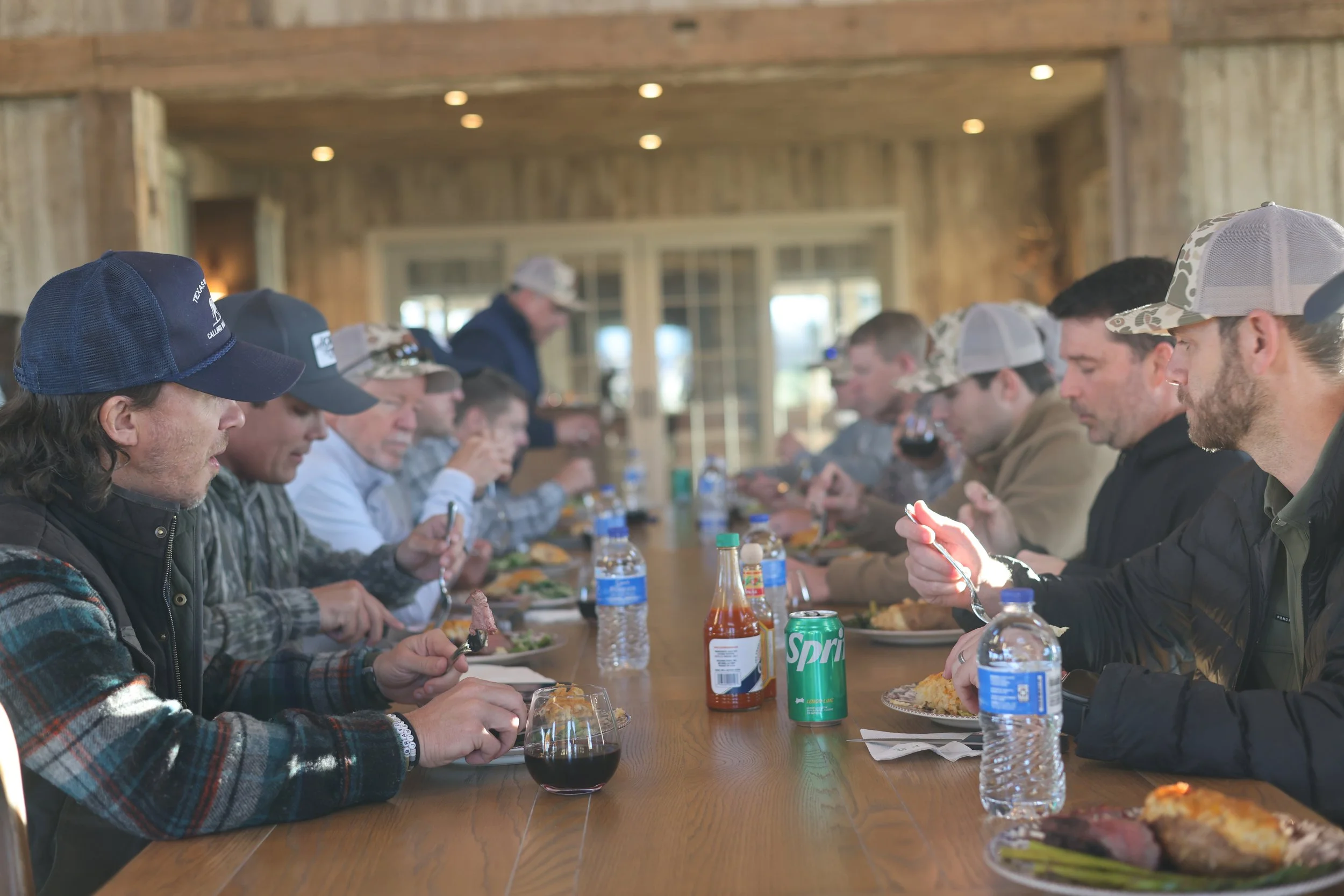 People sitting at a long wooden table enjoying a meal in a rustic room with wooden walls and ceiling, under recessed lighting.
