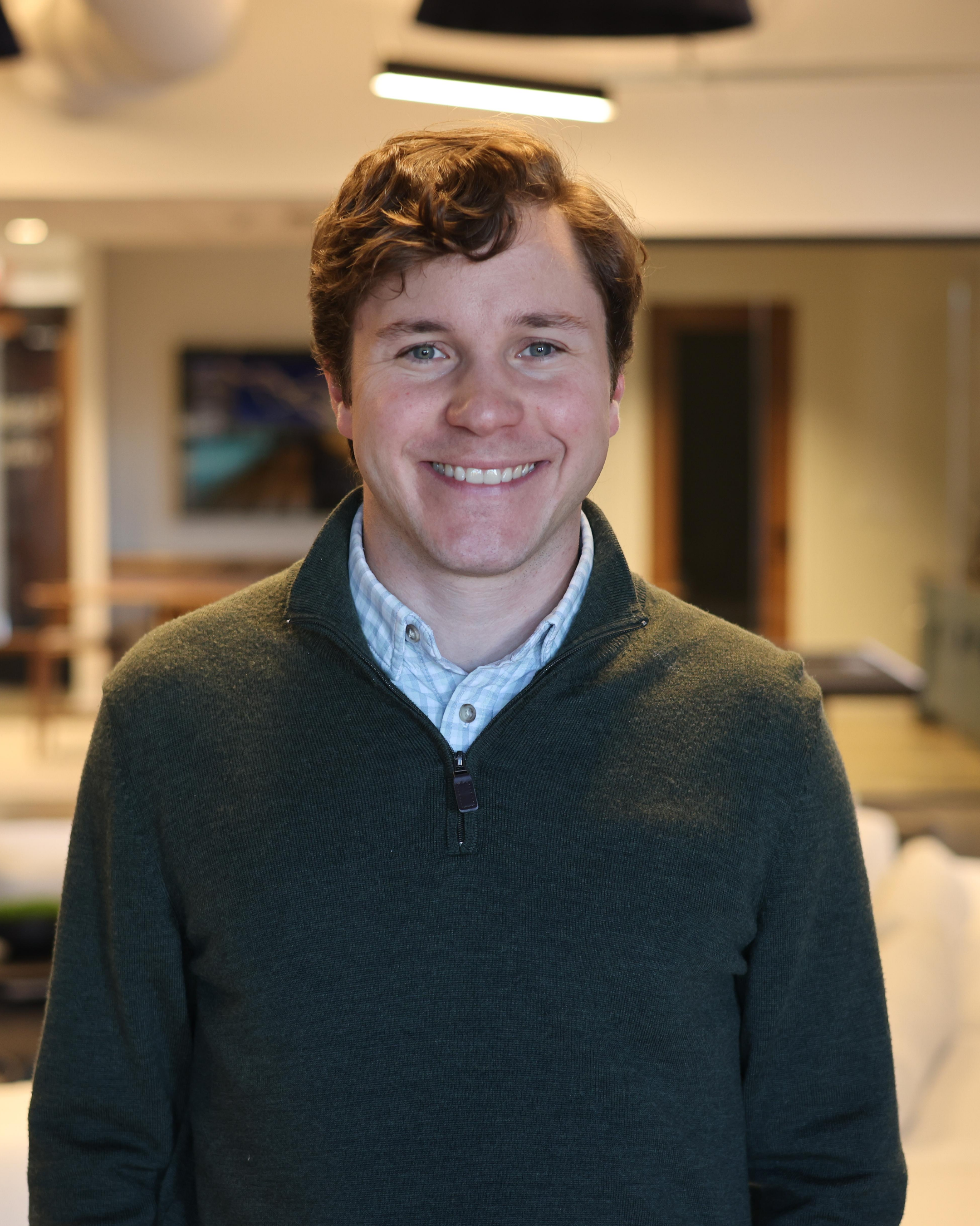 A young man with light skin, curly brown hair, and blue eyes smiling in an indoor setting. He is wearing a dark green zip-up sweater over a button-up shirt. The background shows a blurred interior with a TV and a ceiling light.