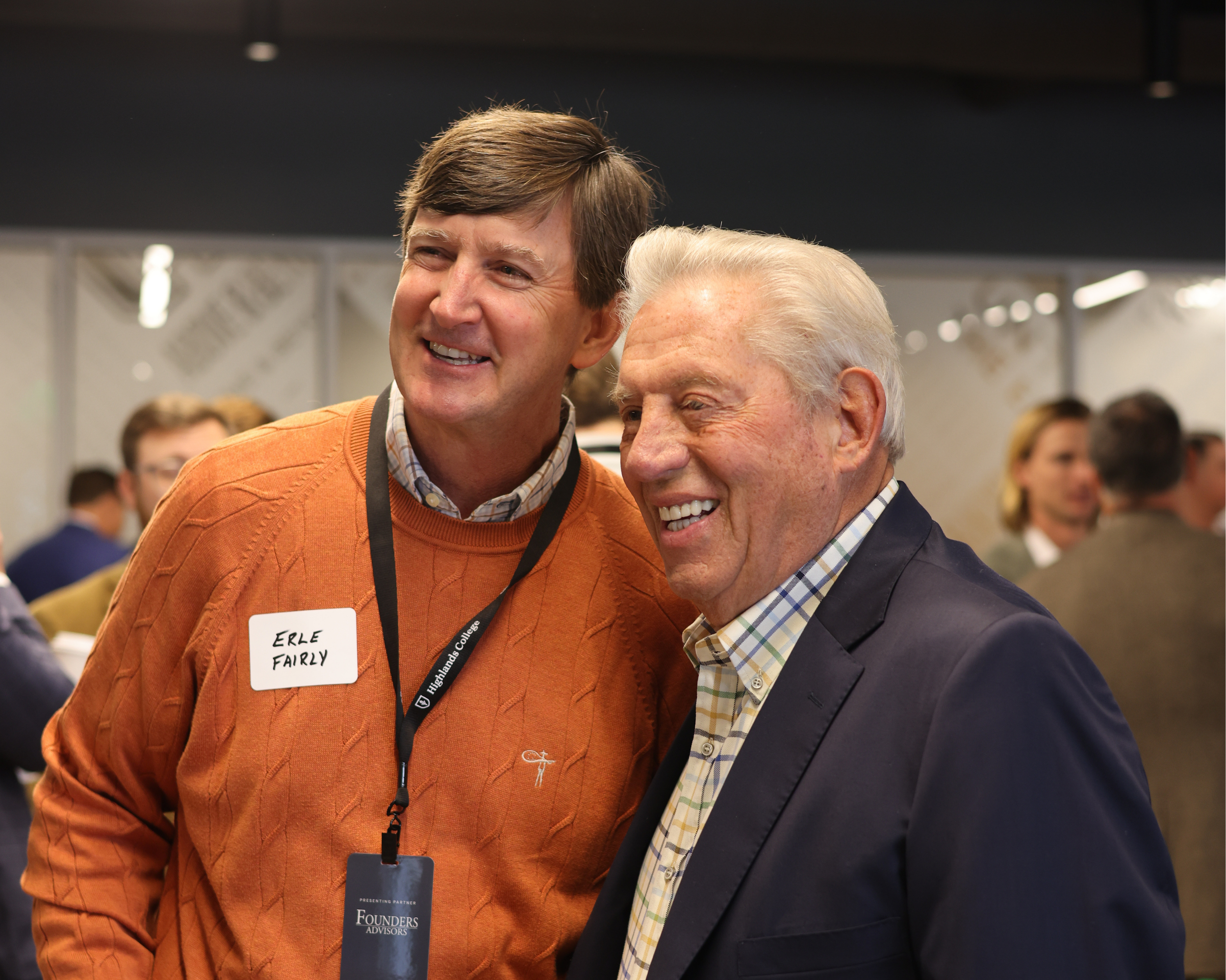 Two men smiling and talking at an indoor event, one wearing an orange sweater with a name tag reading 'Erle Fairly', and the other in a dark blazer and checkered shirt, surrounded by other people in the background.