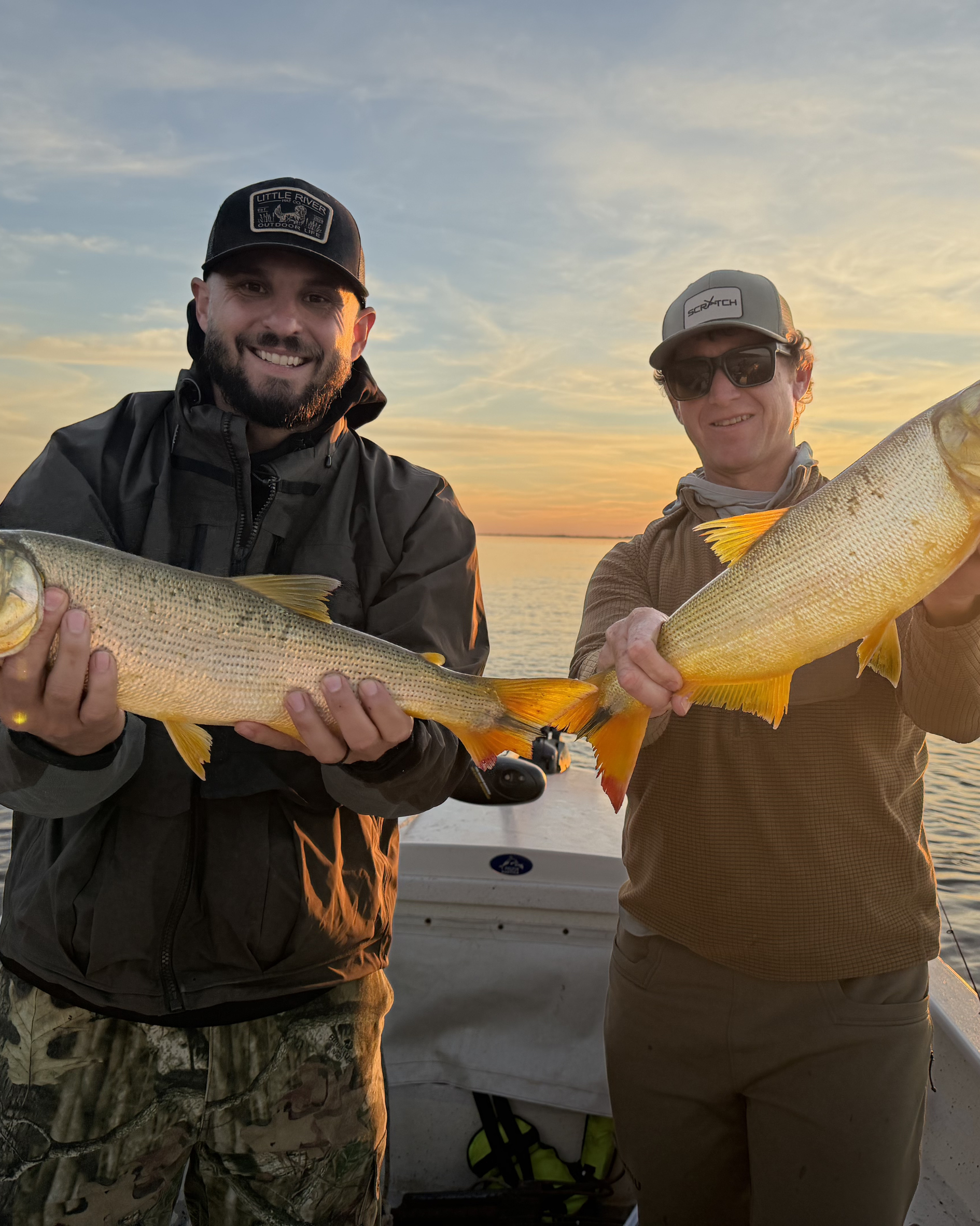 Two men holding large fish on a boat during sunset, wearing outdoor gear and hats.