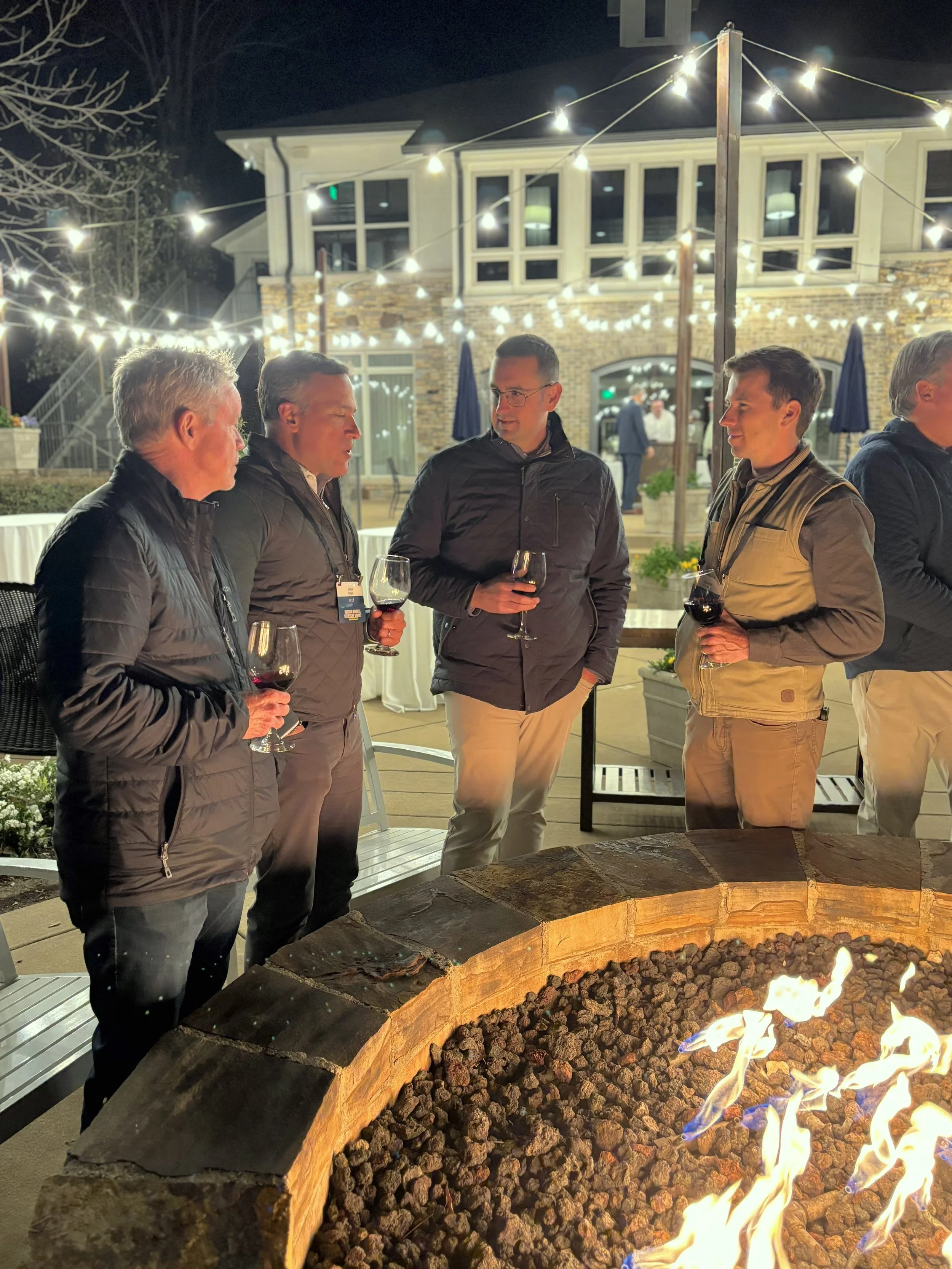 A group of five men standing around a fire pit at night, holding glasses of red wine, engaged in conversation, with string lights and a brick house in the background.