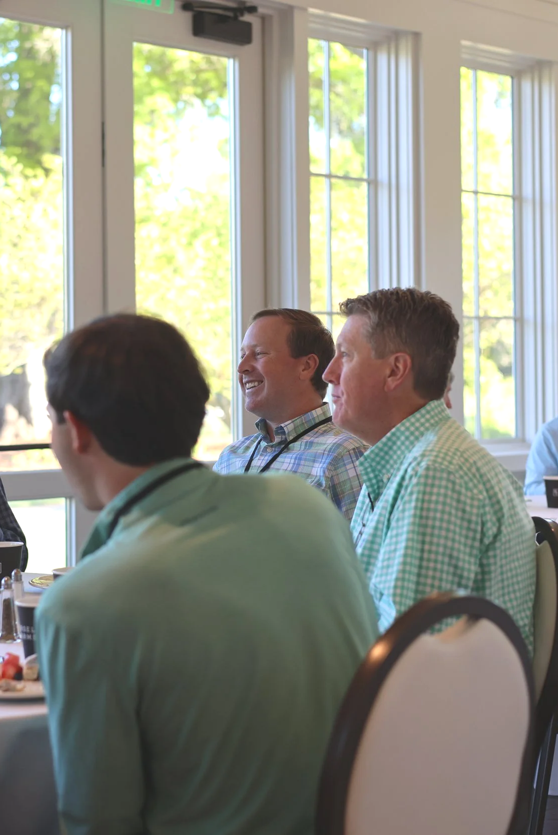 People sitting at a table during a meeting or conference, smiling and engaged, with large windows showing trees outside.