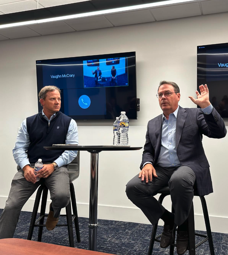 Two men are seated on stools during a panel discussion in a conference room. One man, on the right, is speaking and raising his hand, while the other man, on the left, listens and holds a water bottle. A small round table with two water bottles is between them. Behind them, a large screen displays their image and the name Vaughn McCrary.