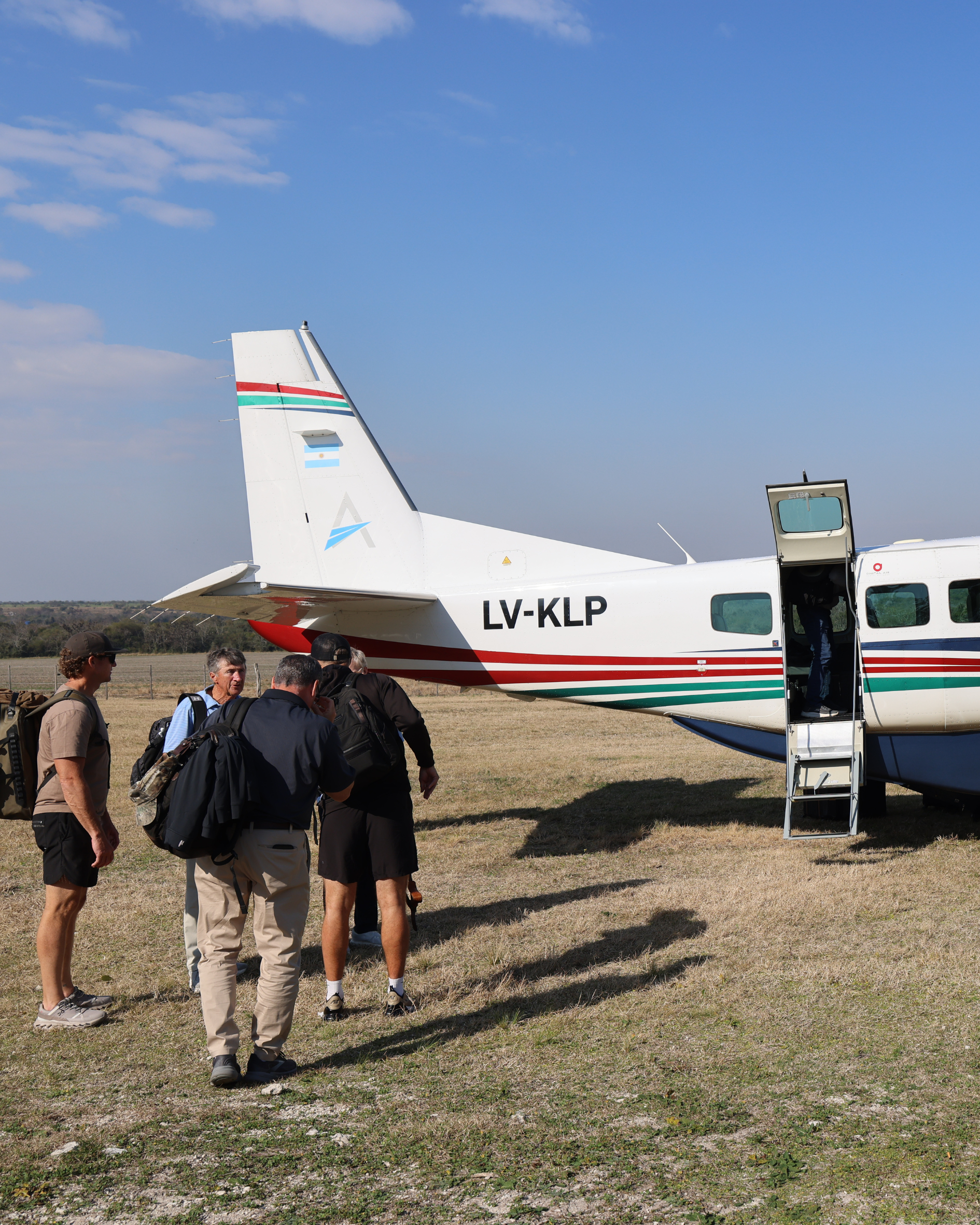 A small group of people walking toward a parked small aircraft on a grassy field with some trees in the background. The aircraft has a white body, with the tail number LV-KLP, and an Argentine flag painted on its tail.