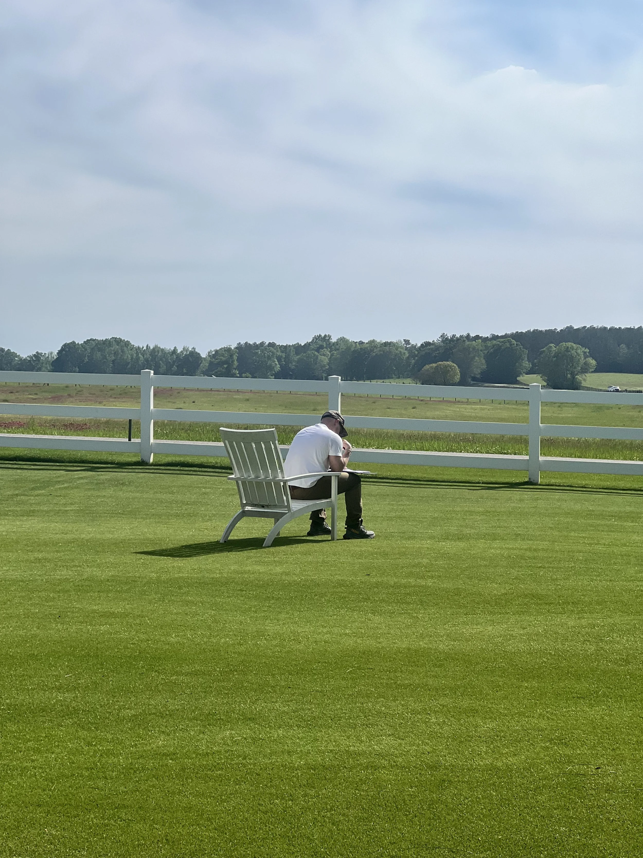 Person sitting alone on a white chair in a grassy outdoor area with a white fence, trees, and a cloudy sky in the background.