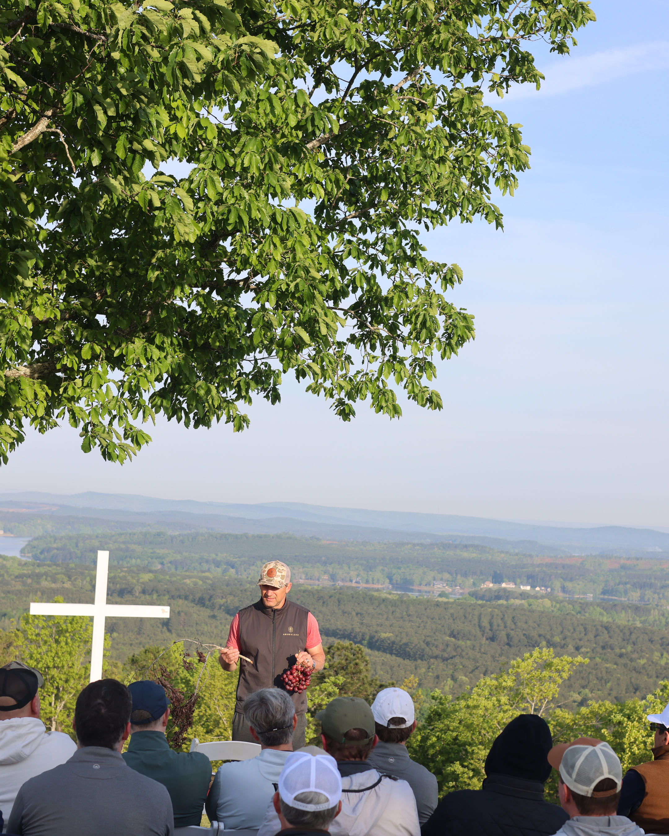 A man in a baseball cap and vest is leading a wine grape harvest presentation outdoors to a group of people sitting under a tree, with rolling hills and blue sky in the background.